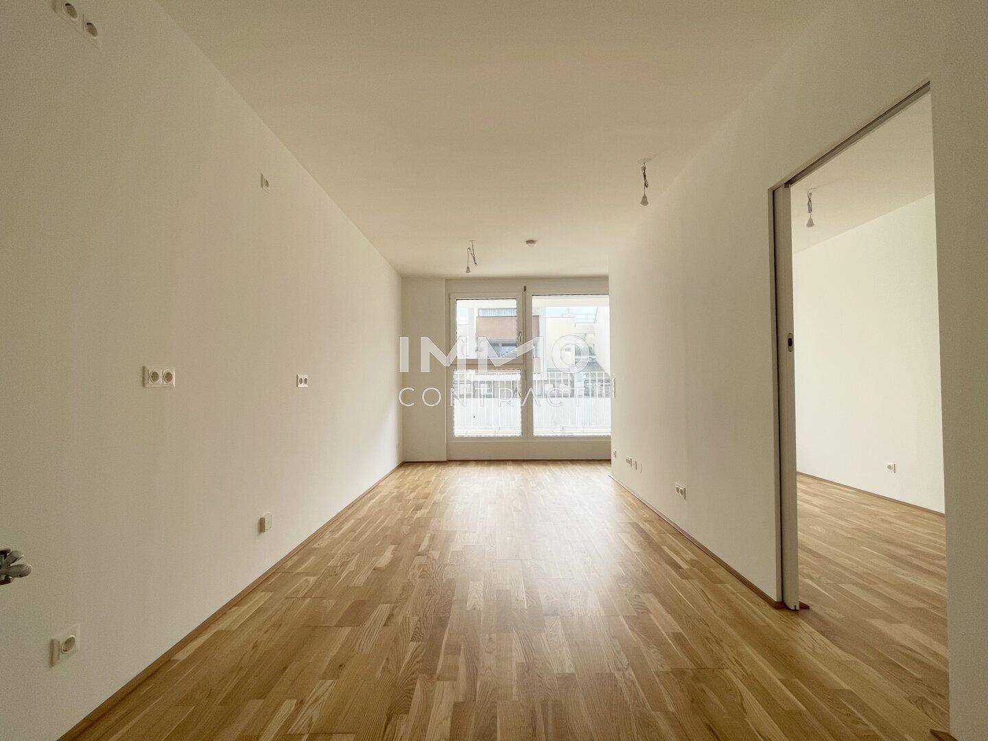 Heller, leerer Wohnraum mit Holzboden und großen Fenstern zum Balkon.