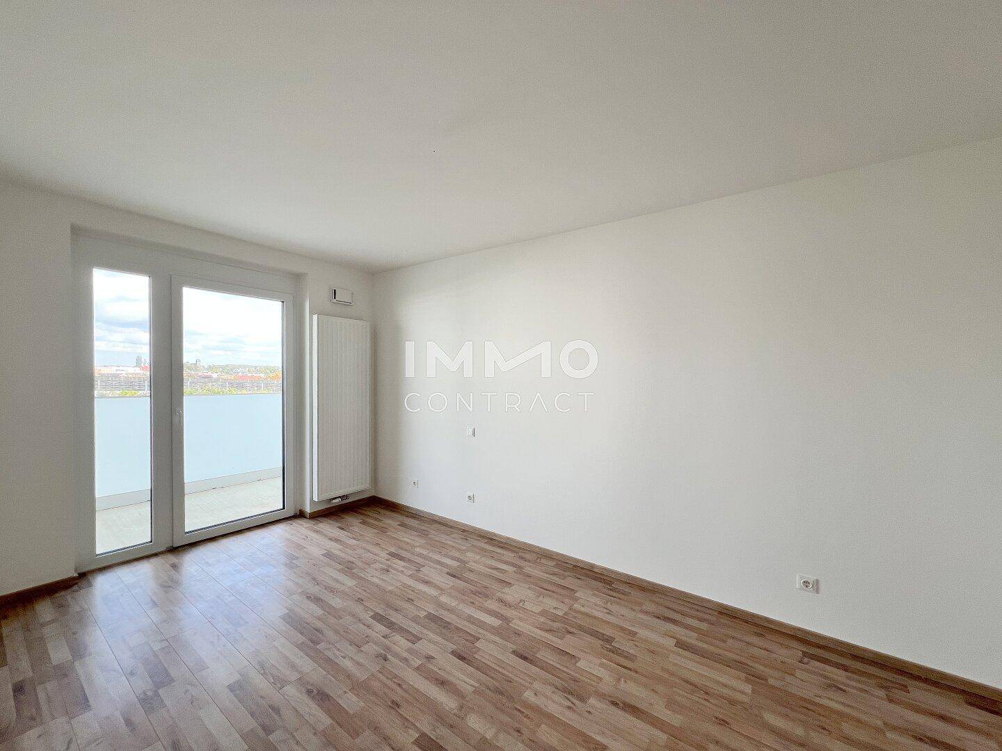 Leeres Schlafzimmer mit Laminatboden und großen Fenstern, die Zugang zum Balkon bieten.