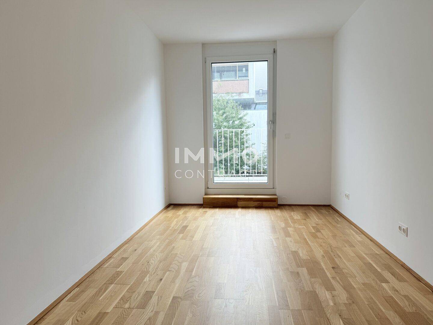 Helles, leeres Schlafzimmer mit Holzboden und Zugang zu einem kleinen Balkon.