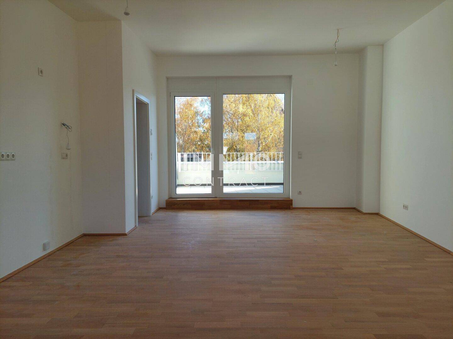 Heller, leerer Raum mit Holzboden und Zugang zu einem Balkon mit Blick ins Grüne.