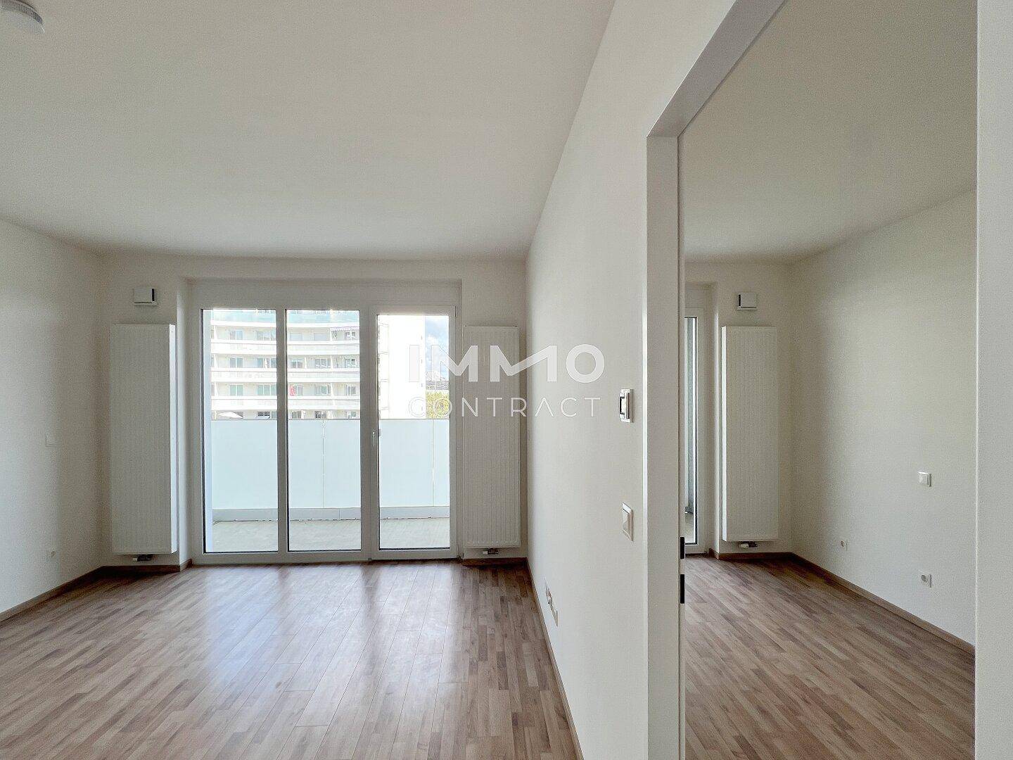 Helles, leeres Wohnzimmer mit Holzboden und großen Schiebetüren zum Balkon.