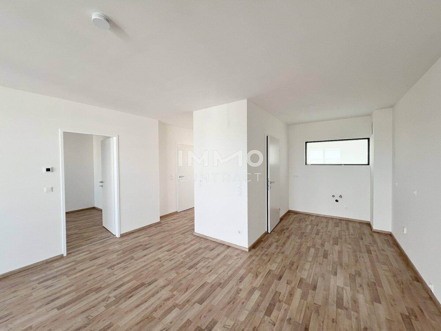 Helles Wohnzimmer mit Laminatboden und vorbereiteten Anschlüssen für eine Küche.