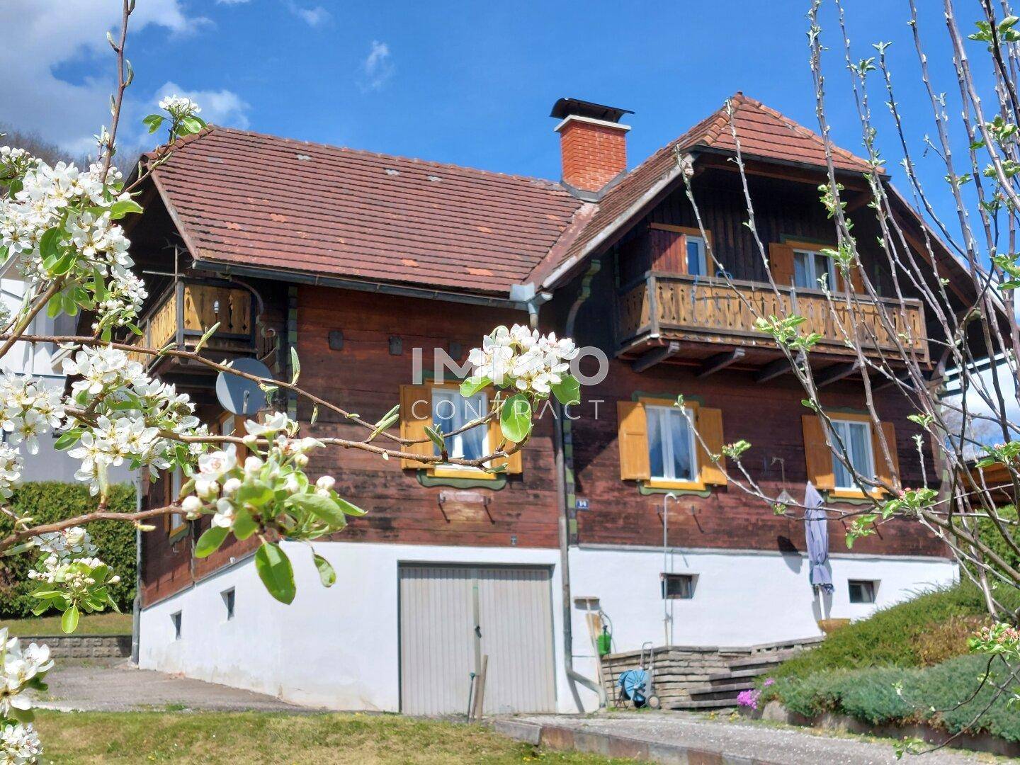 Charmantes Holzhaus mit blühenden Bäumen im Vordergrund, umgeben von grüner Natur.