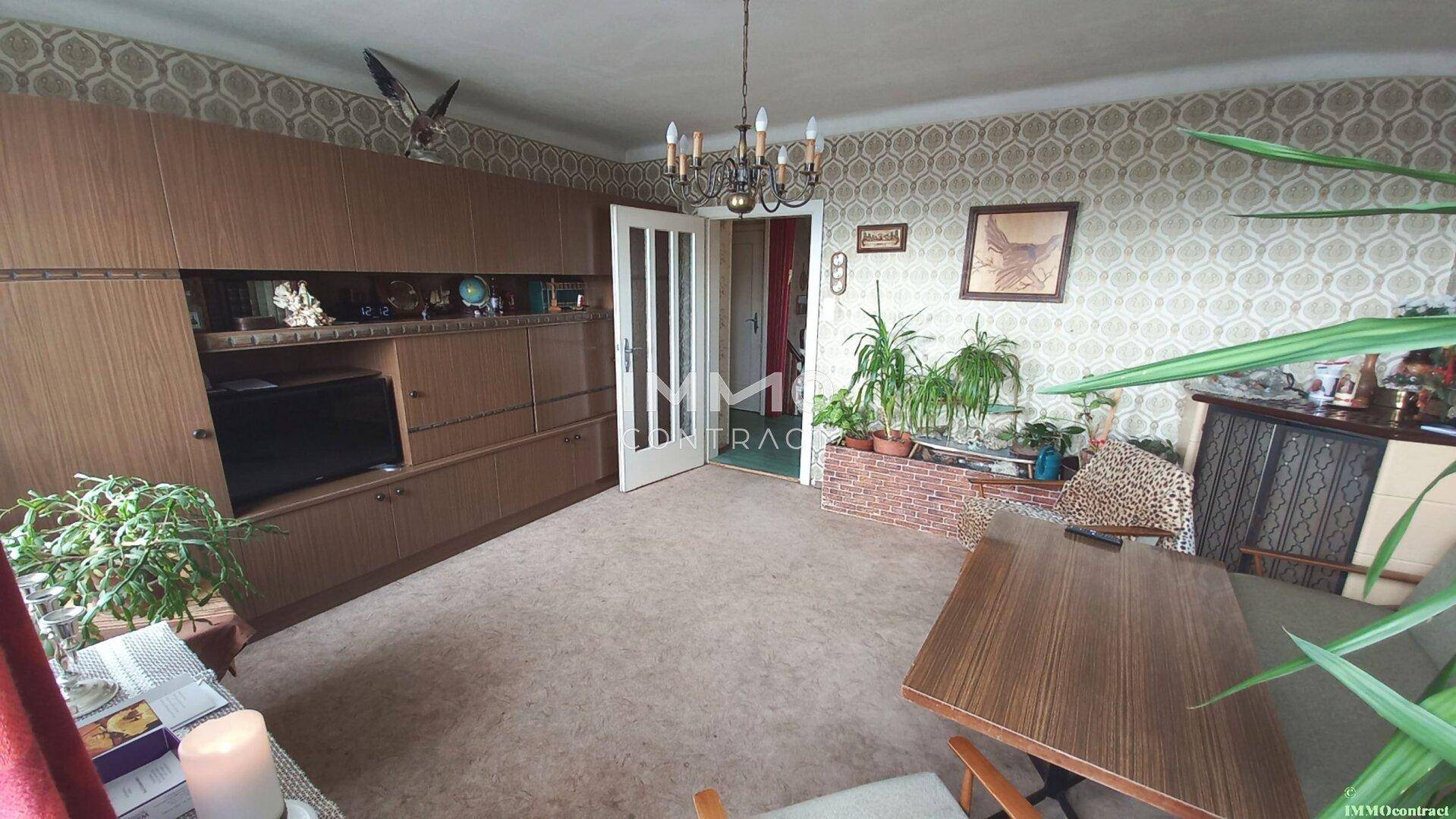 Geräumiges Wohnzimmer mit Teppichboden, großer Schrankwand und Pflanzen.