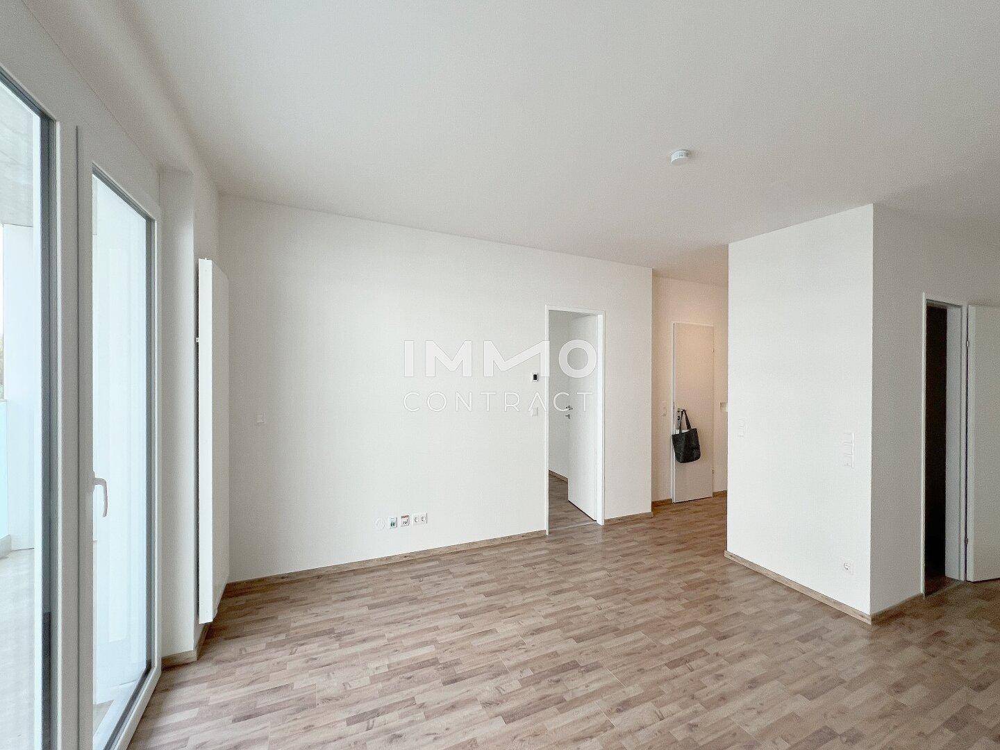 Geräumiges, leeres Wohnzimmer mit Laminatboden und großen Glasfenstern zum Balkon.