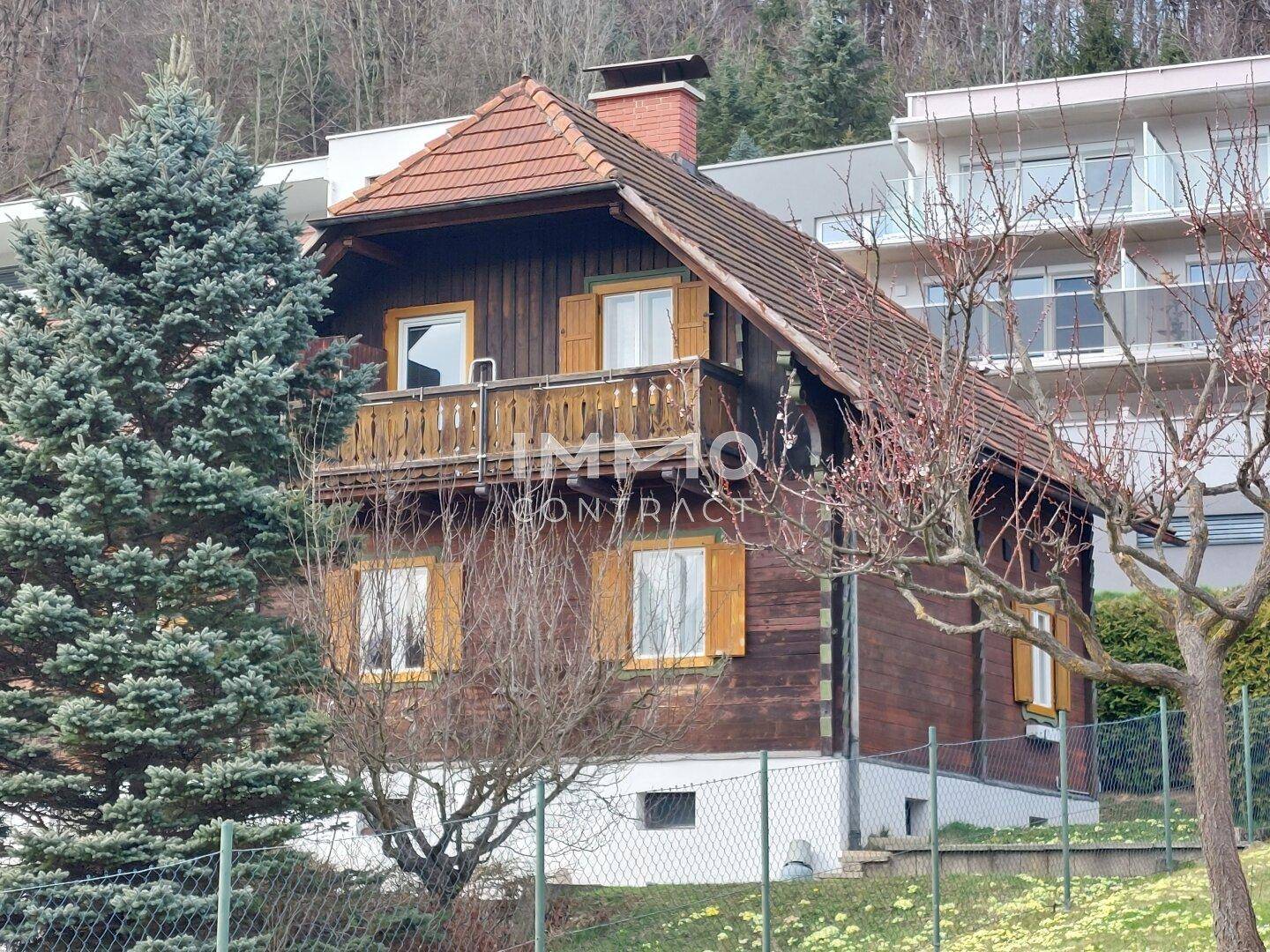 Traditionelles Holzhaus mit Satteldach, umgeben von Bäumen und modernen Gebäuden im Hintergrund.