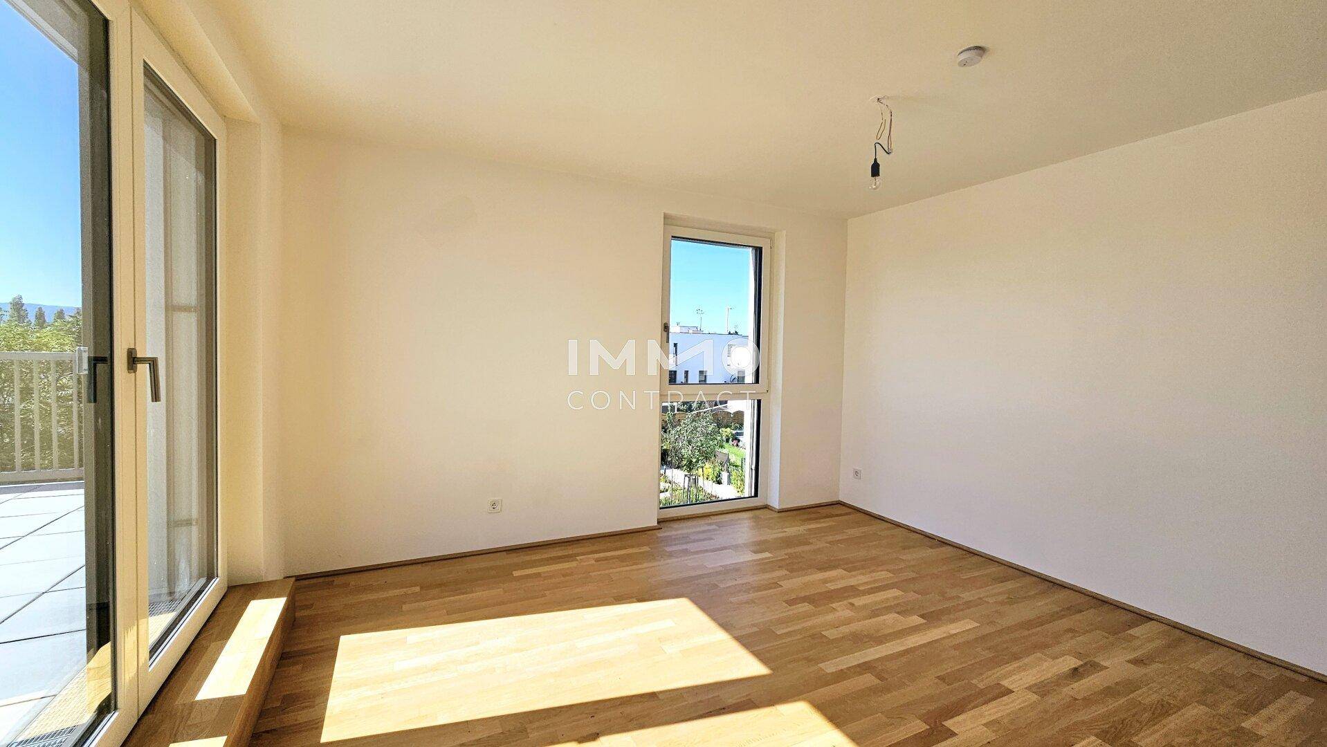 Helles Zimmer mit Holzboden und großem Fenster, das viel Tageslicht hereinlässt und einen Blick ins Grüne bietet.