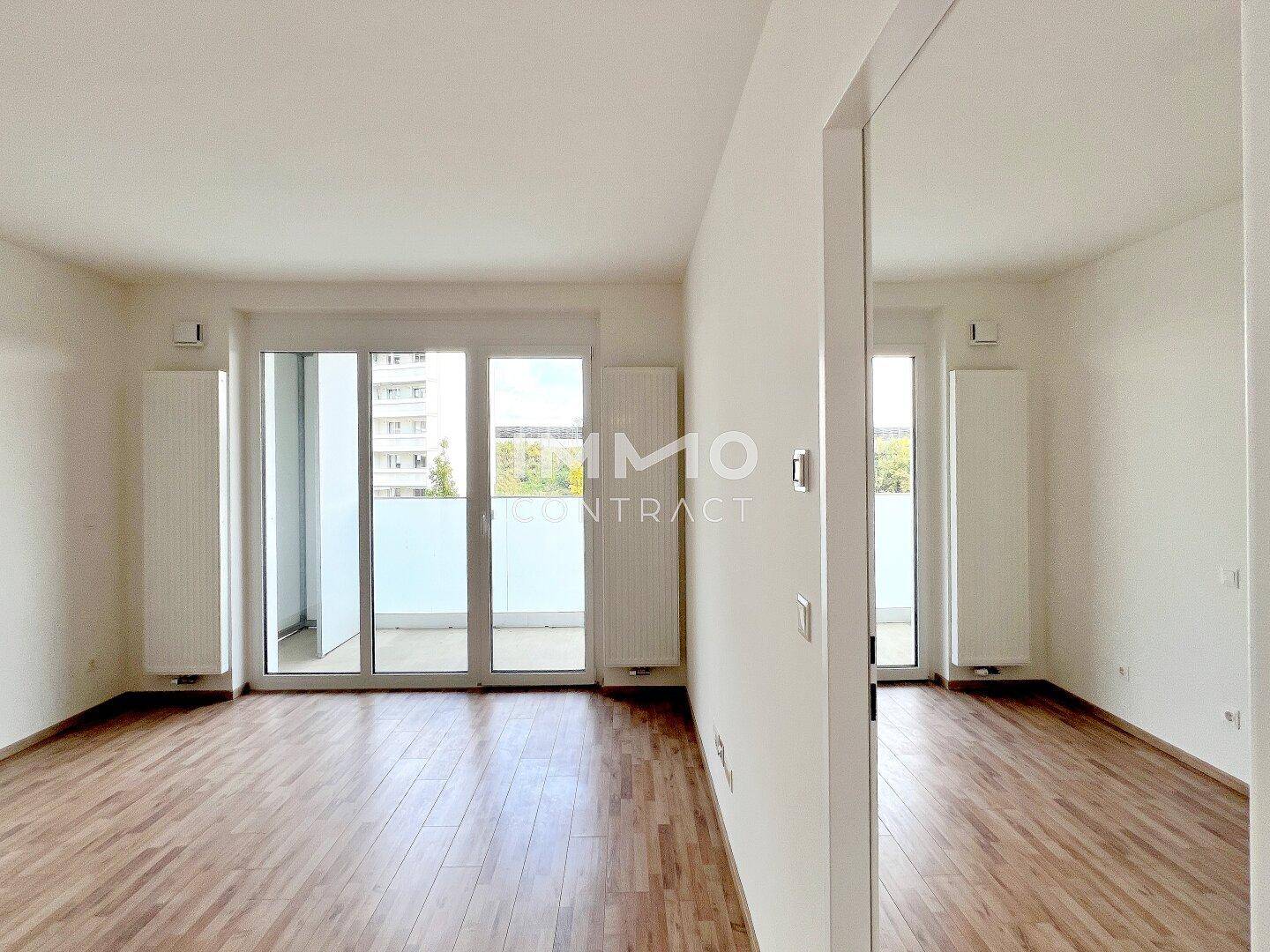 Leeres Wohnzimmer mit hellem Laminatboden, großen Fenstern und direktem Zugang zum Balkon.
