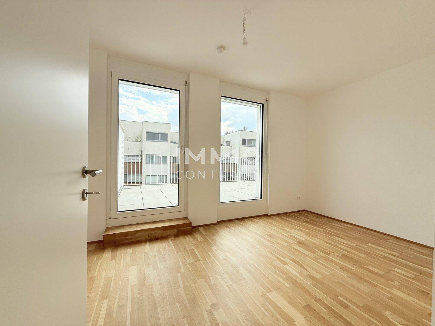 Helles, leeres Zimmer mit Parkettboden und Zugang zur Terrasse durch große Fenster.