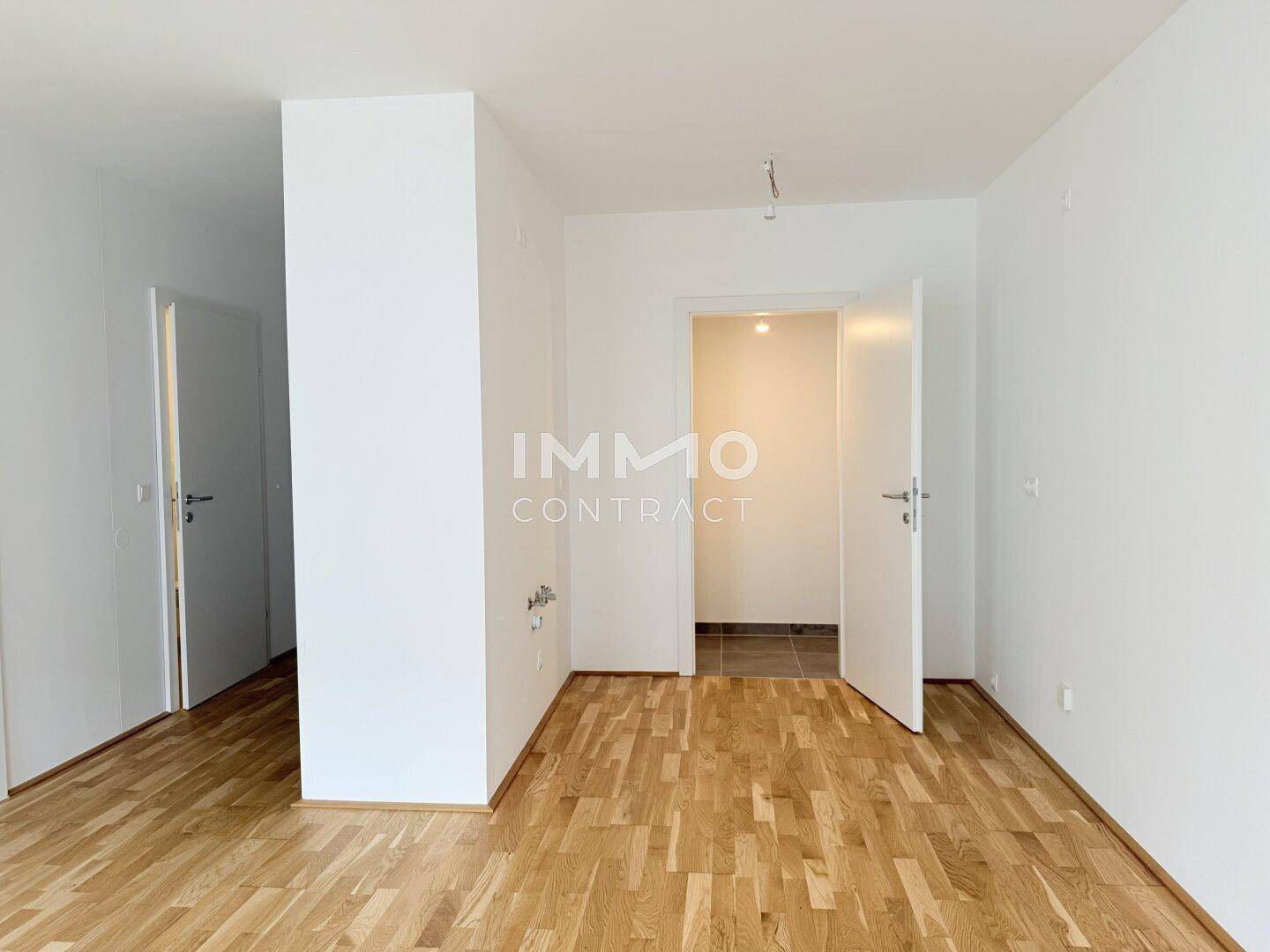 Leerer Innenraum mit Holzboden und weißen Wänden, der zu weiteren Räumen führt.