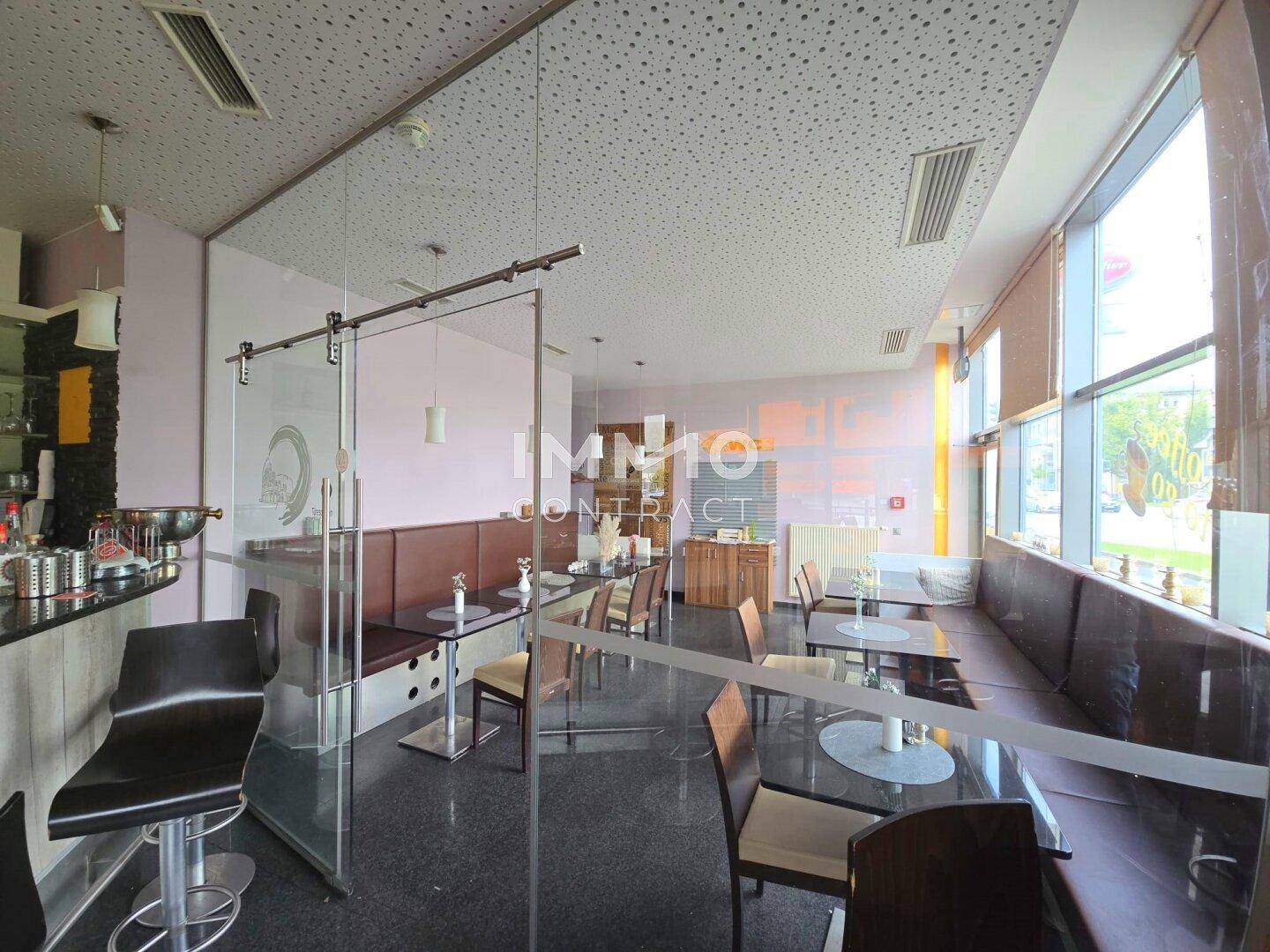 Moderner Café-Innenraum mit Glasabtrennung, bequemen Sitzbänken und großen Fenstern.