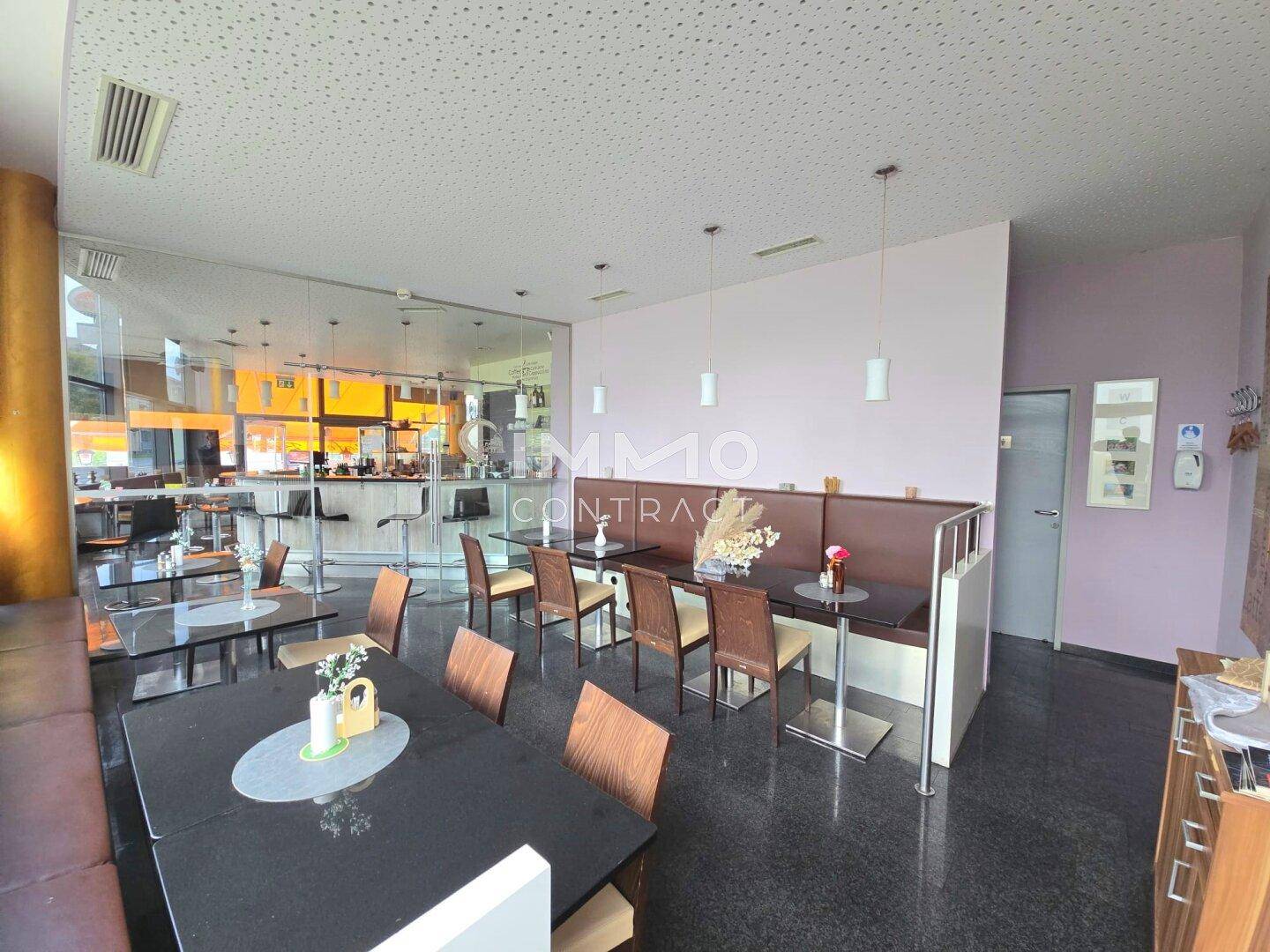 Geräumiger Café-Sitzbereich mit bequemen Bänken, Tischen und Blick auf die Bar durch eine Glaswand.