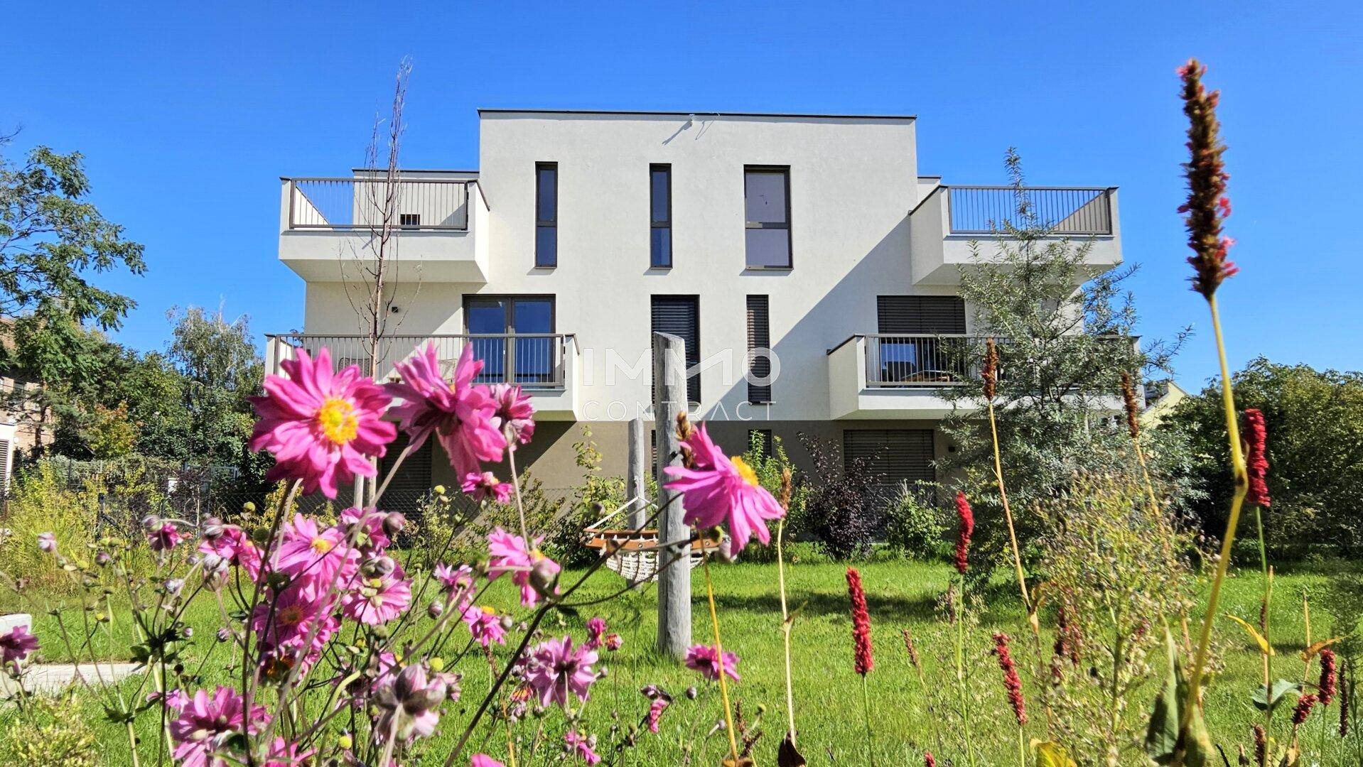 Moderne Wohnanlage mit weißer Fassade und Balkonen, umgeben von blühenden Pflanzen und Grünflächen.