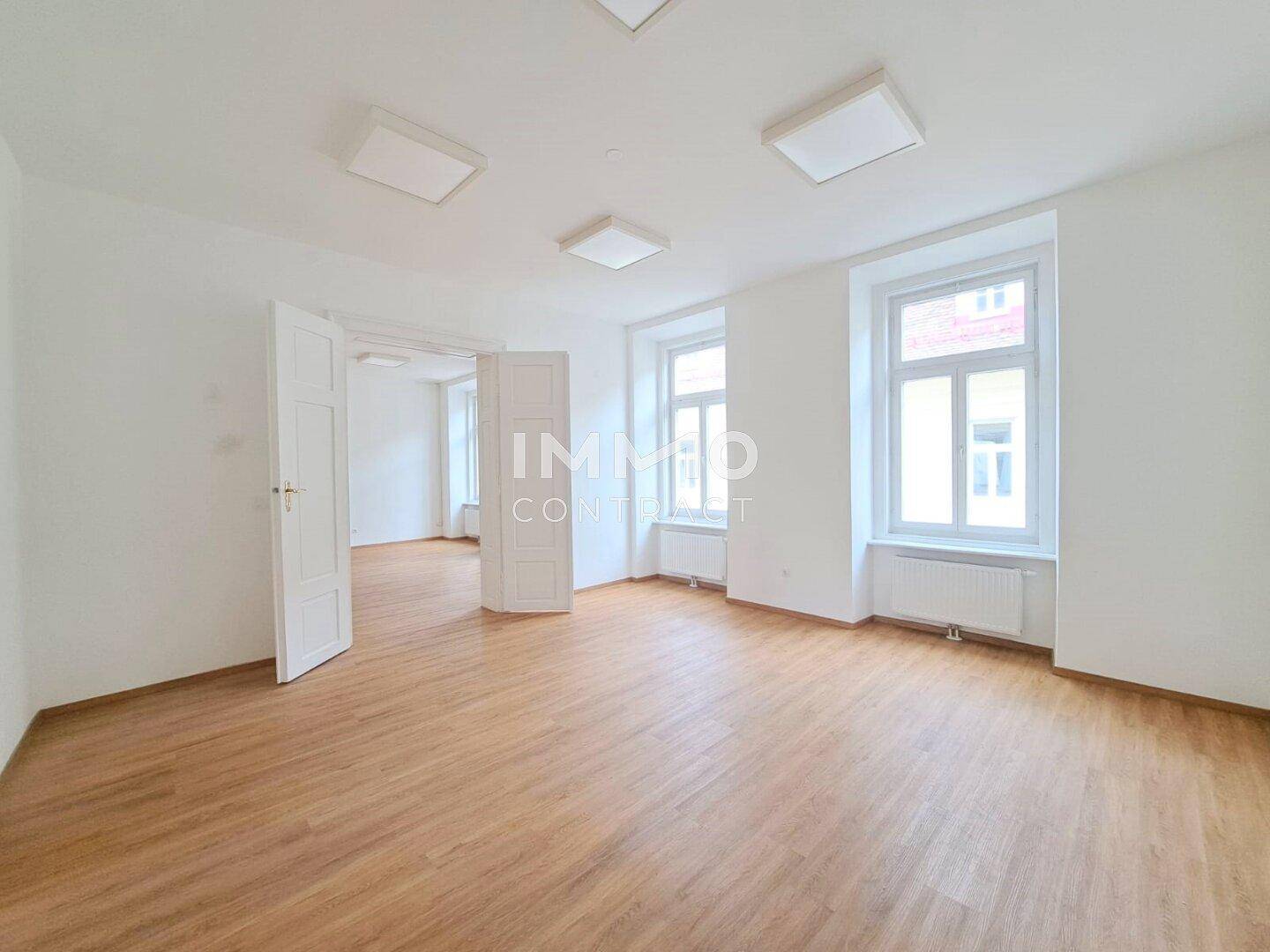 Geräumiger, heller Raum mit Holzboden und Doppeltür, der in einen weiteren Bereich führt.