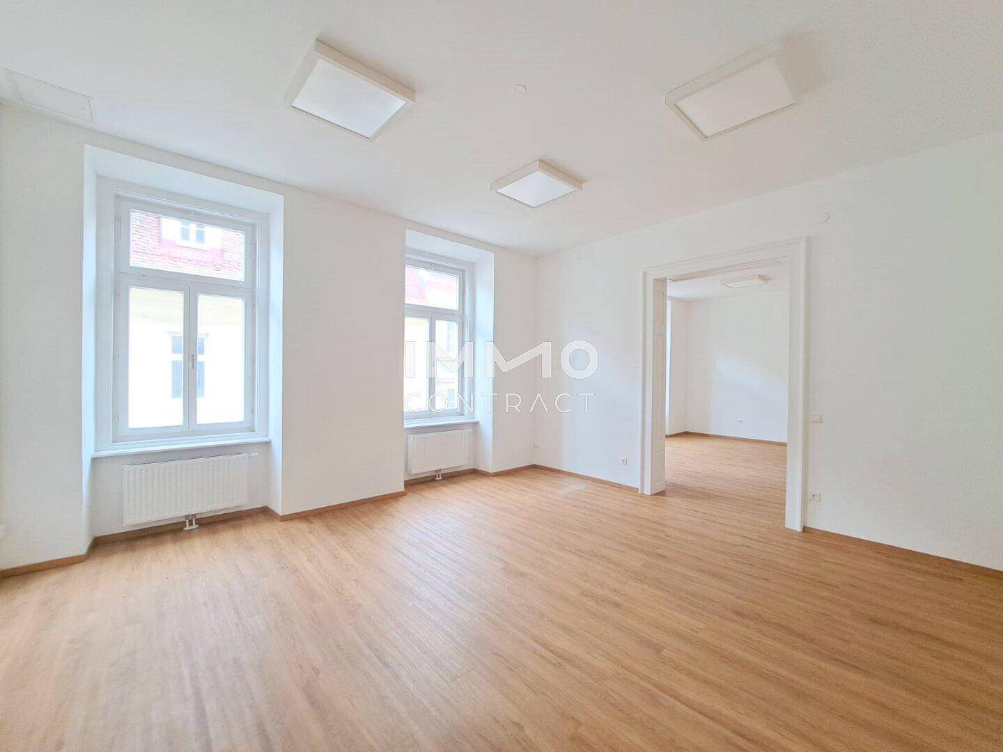 Heller, leerer Raum mit Holzboden und zwei großen Fenstern, ideal für Wohn- oder Bürozwecke.