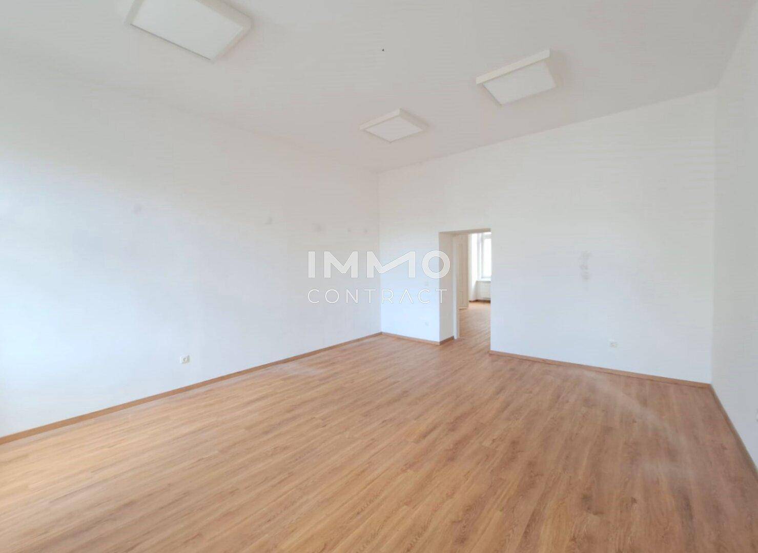 Geräumiger, heller Raum mit Holzboden und Zugang zu einem weiteren Zimmer, mit viel Tageslicht.