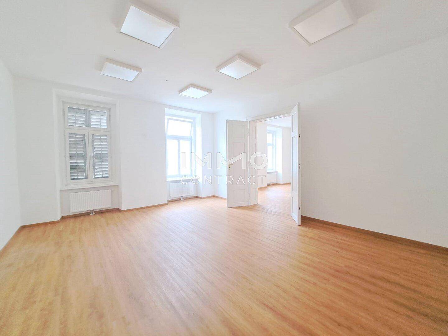 Geräumiger, heller Raum mit Holzboden und Doppeltür, der in einen weiteren Bereich führt, mit Fensterläden.
