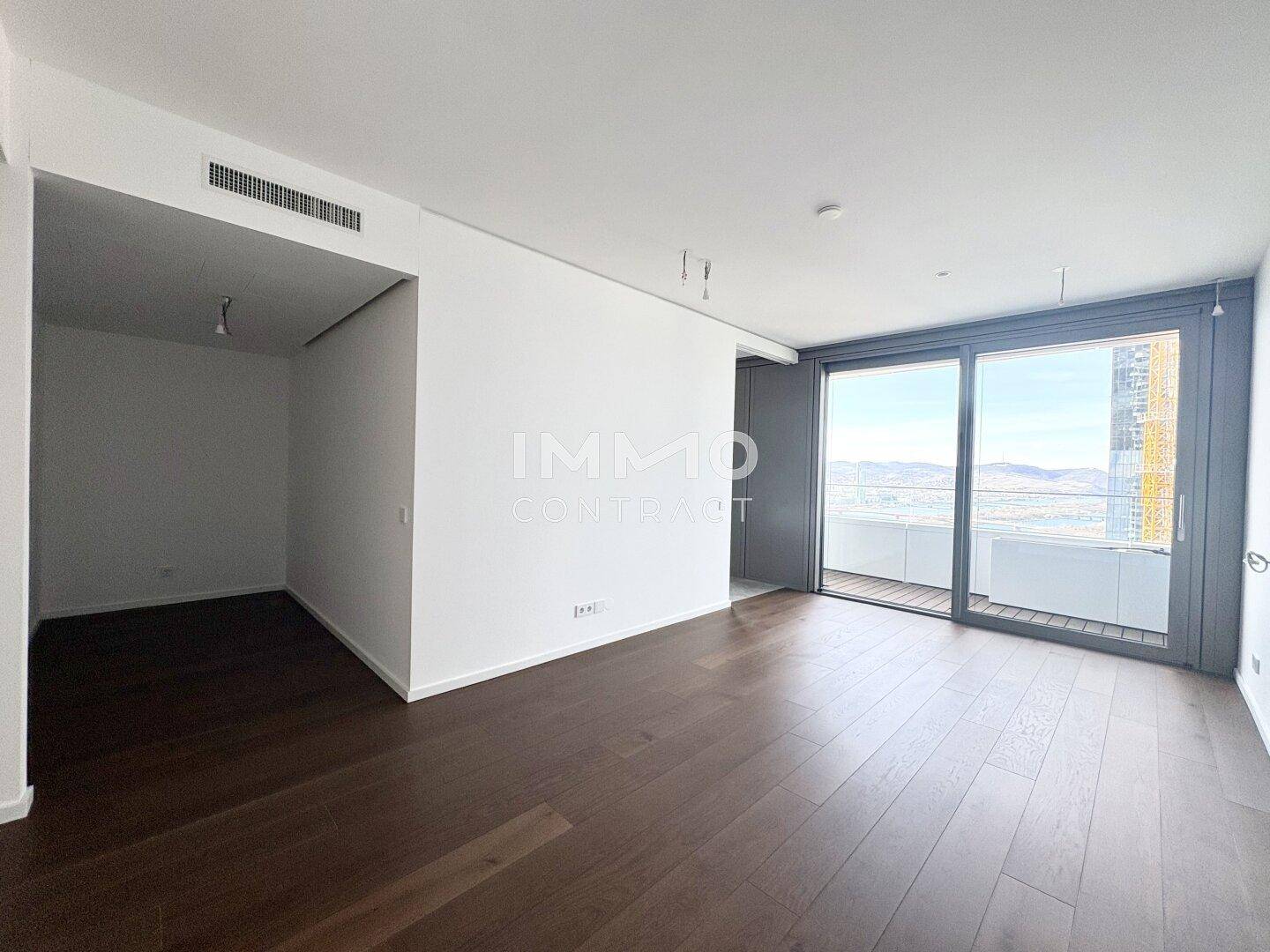 Heller, leerer Raum mit dunklem Holzboden und Zugang zu einem Balkon mit Stadtblick.