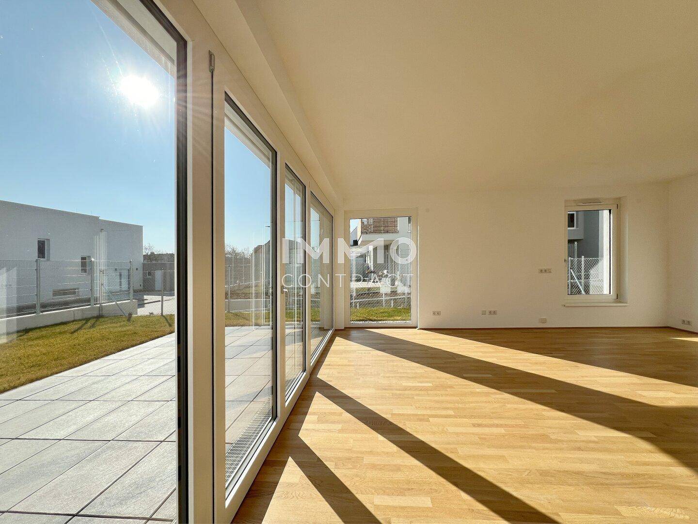 Heller, leerer Wohnbereich mit großen Fenstern, Parkettboden und Zugang zur Terrasse.