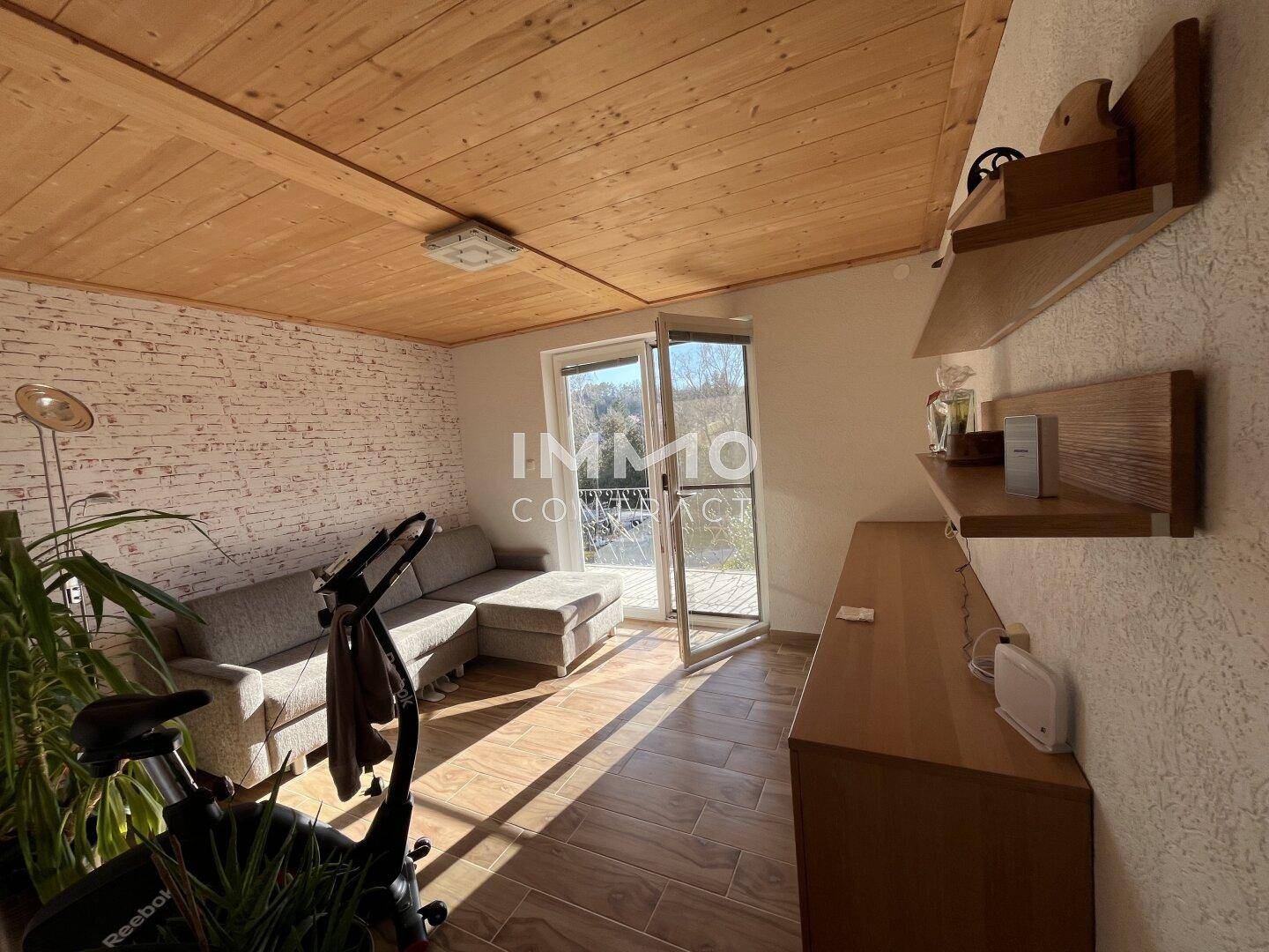 Gemütliches Wohnzimmer mit Holzdecke, Fliesenboden und Zugang zu einem Balkon.