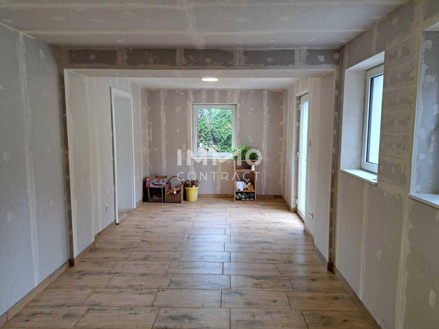 Unfertiger Raum mit Holzoptik-Fliesenboden und Fenstern, bereit für den Ausbau.