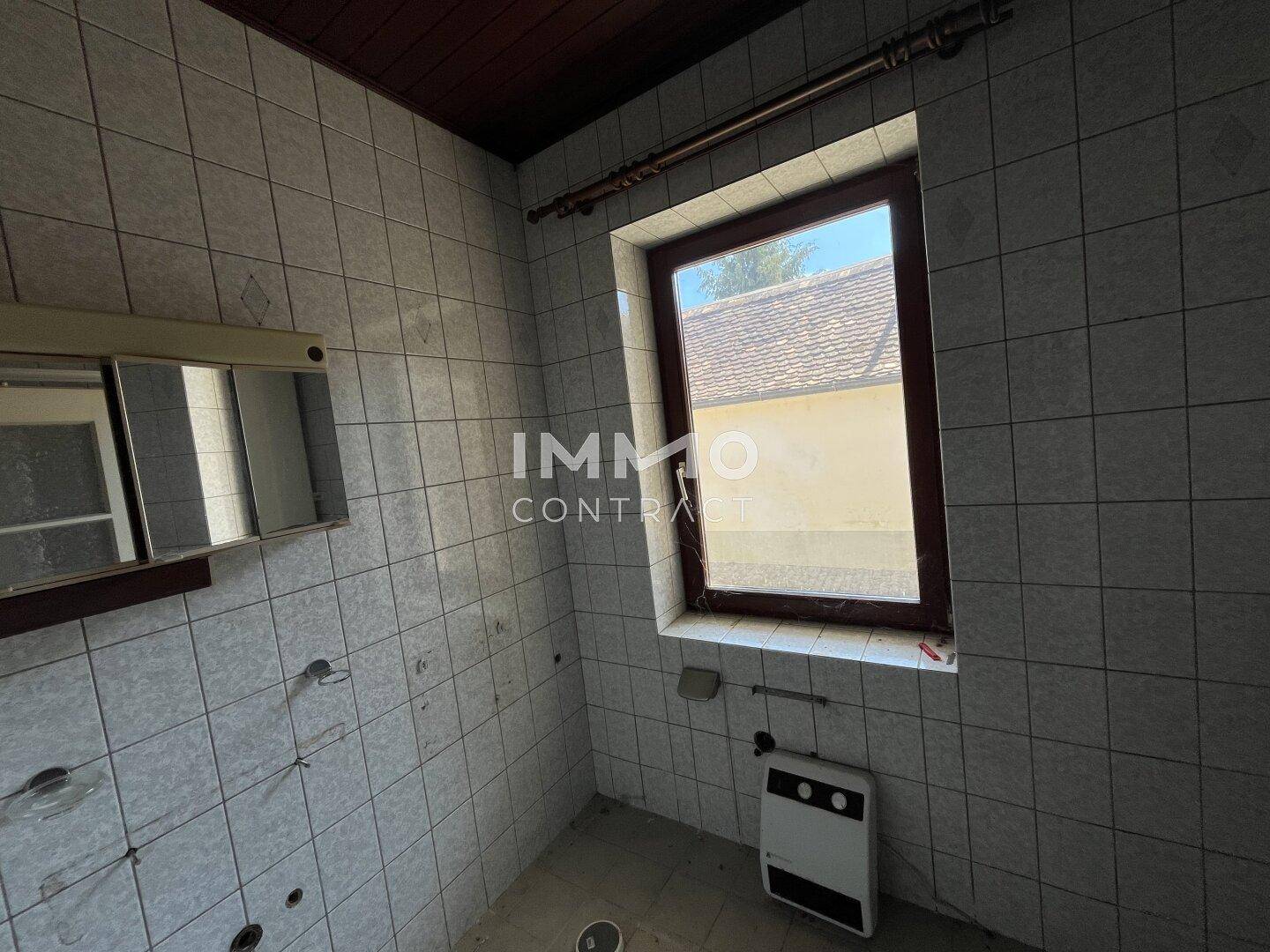 Gefliestes Badezimmer mit Fenster, Spiegelschrank und sichtbaren Anschlüssen für Sanitäranlagen.