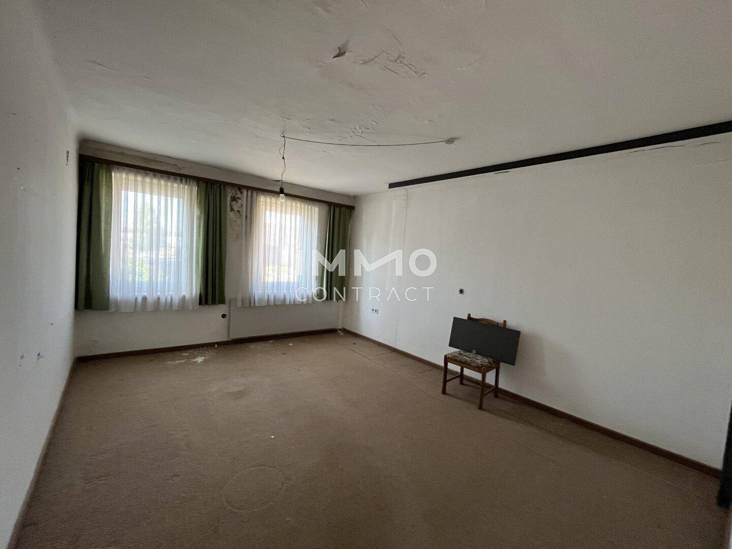 Geräumiges Zimmer mit Teppichboden, zwei Fenstern und sichtbaren Gebrauchsspuren an Wänden und Decke.