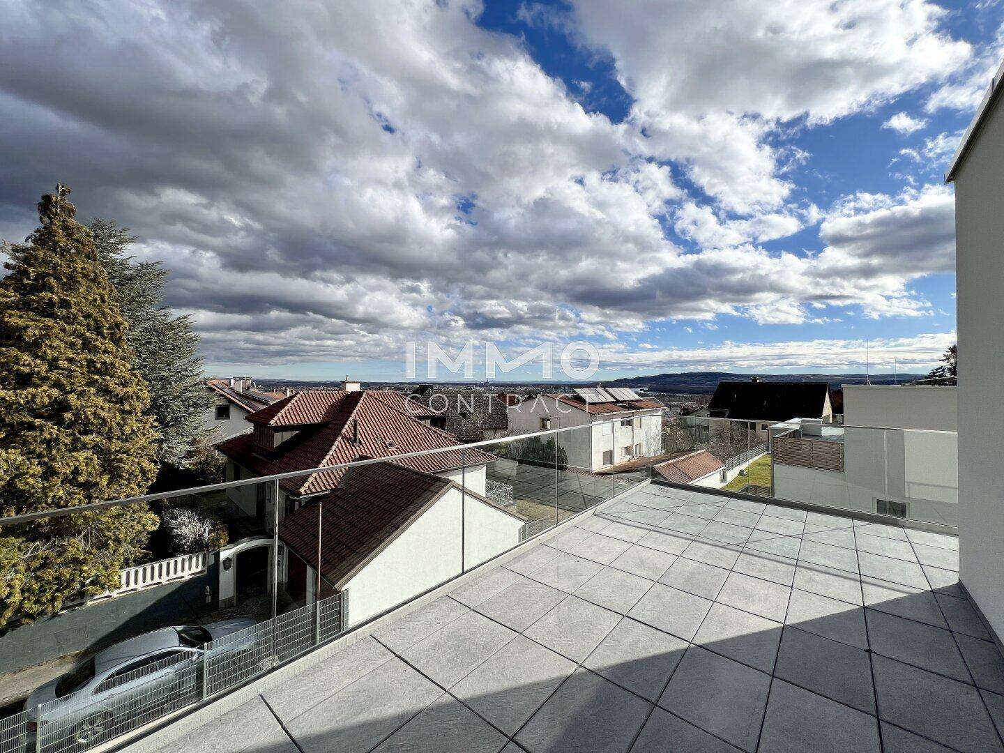 Panoramablick von der Terrasse über eine Wohnsiedlung mit Dächern, Bäumen und hügeliger Landschaft.