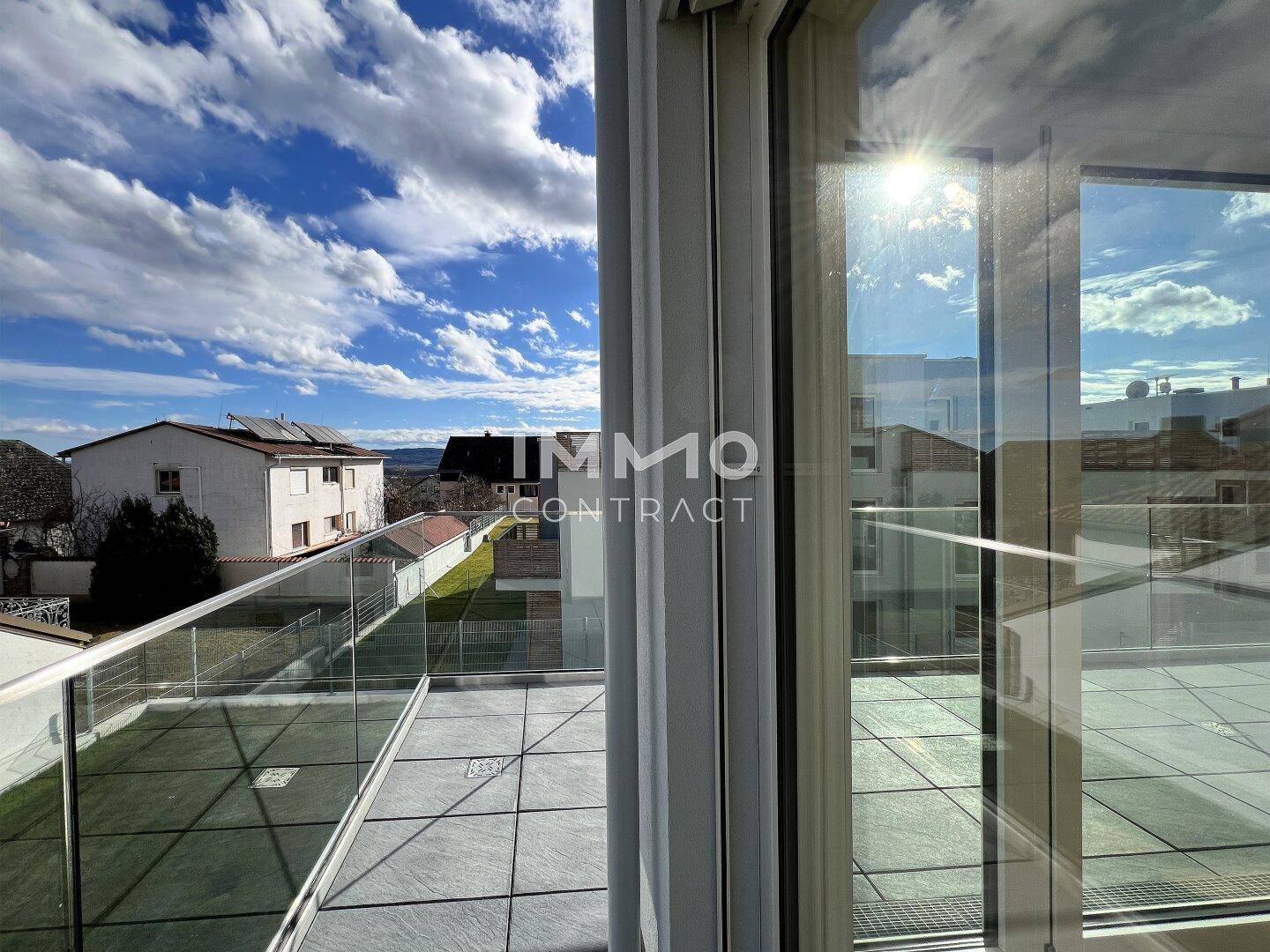 Blick von innen auf die sonnige Terrasse mit Glasgeländer und die umliegende Wohnbebauung.