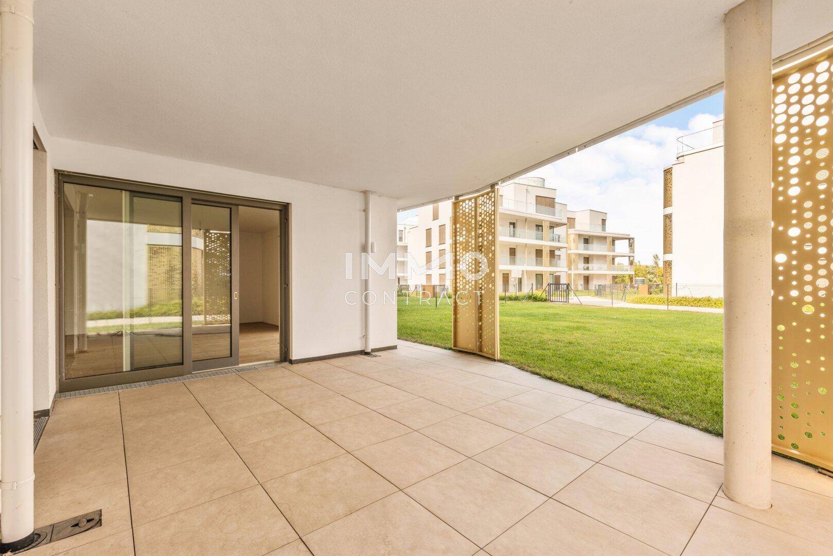 Überdachte Terrasse mit Fliesenboden und Blick auf den Rasen sowie moderne Wohngebäude.