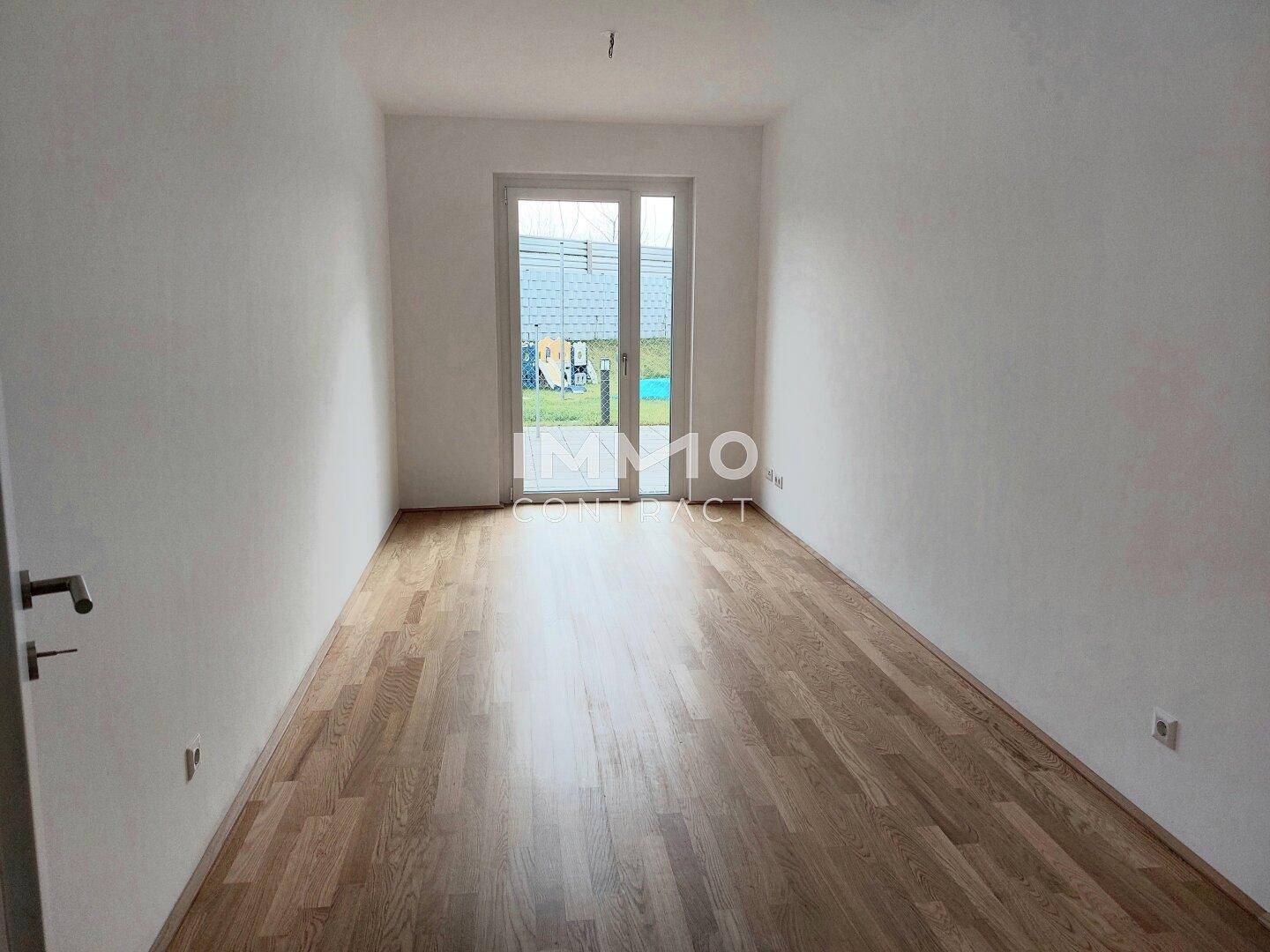 Leeres Zimmer mit Parkettboden und großer Fenstertür, die Zugang zu einem Außenbereich bietet.