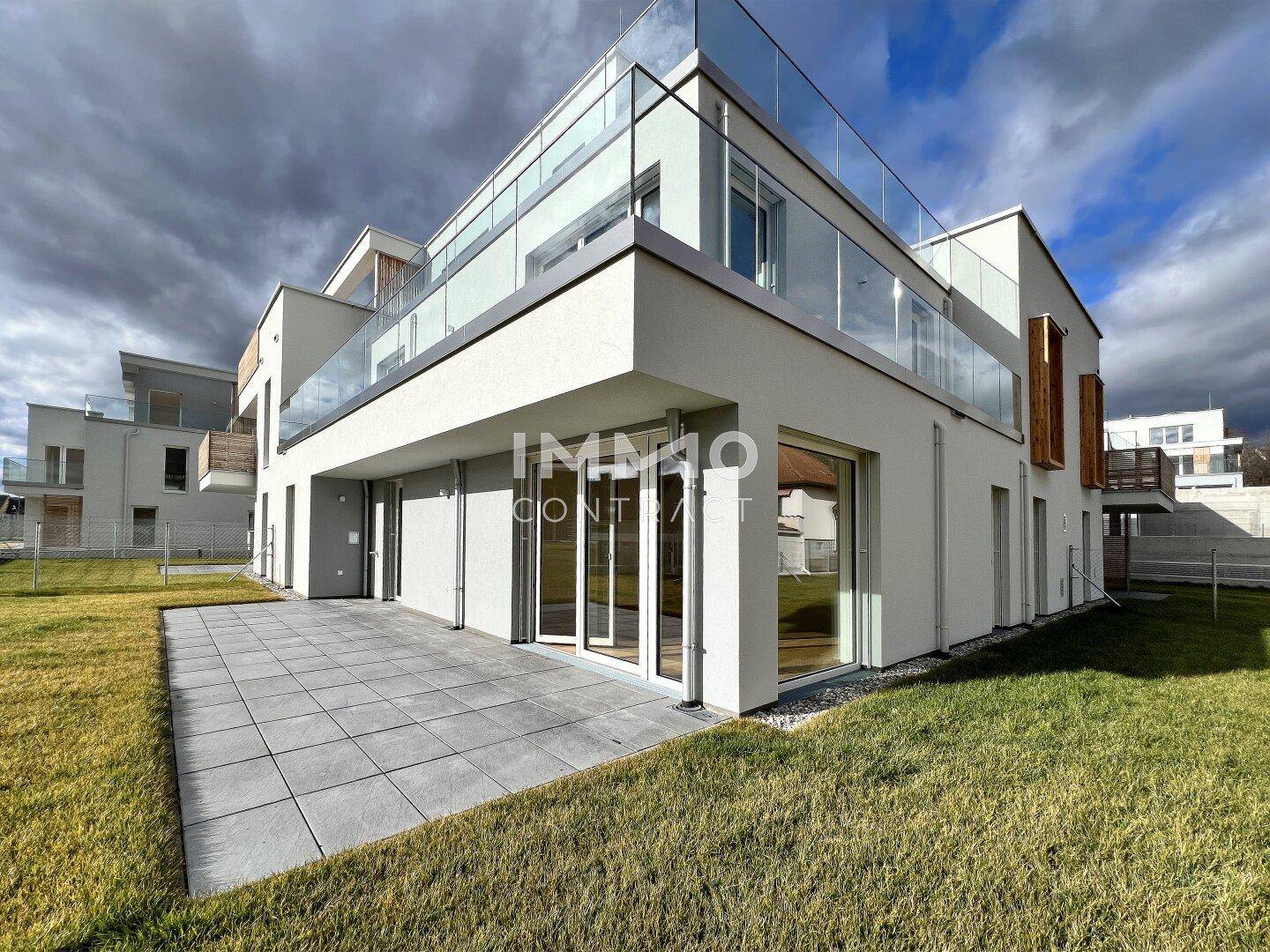 Eckansicht des modernen Gebäudes mit großer gepflasterter Terrasse und angrenzender Rasenfläche unter bewölktem Himmel.