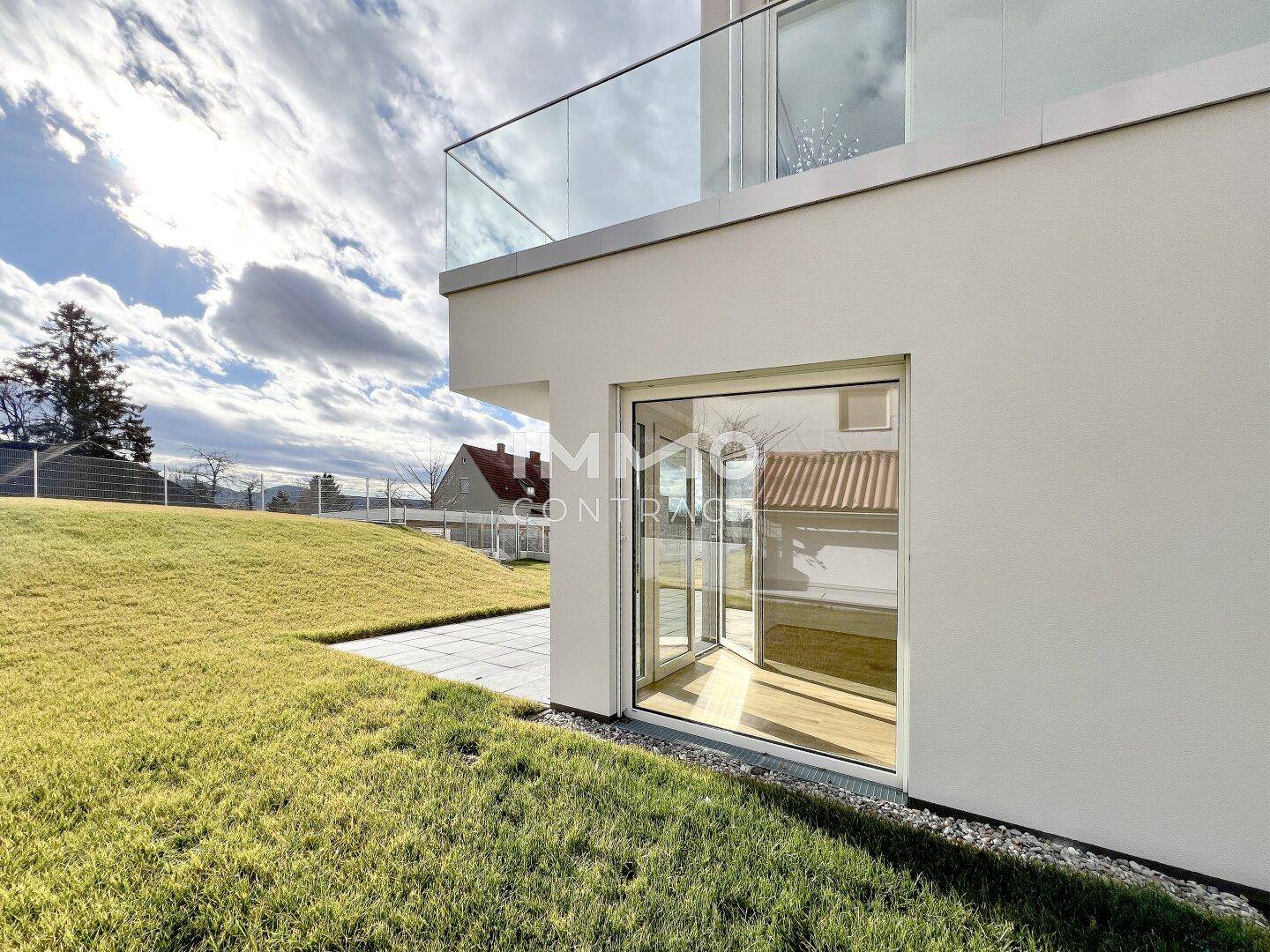 Große Glasfront führt auf eine gepflasterte Terrasse und eine weitläufige Rasenfläche mit Blick in die Ferne.