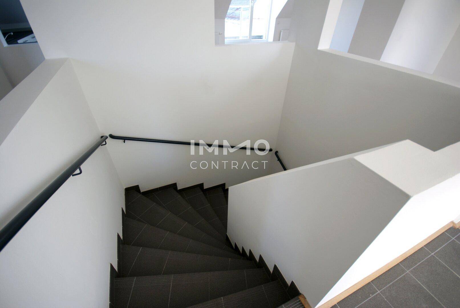 Geräumiges Treppenhaus mit dunklen Fliesenstufen und weiß gestrichenen Wänden, gut beleuchtet.