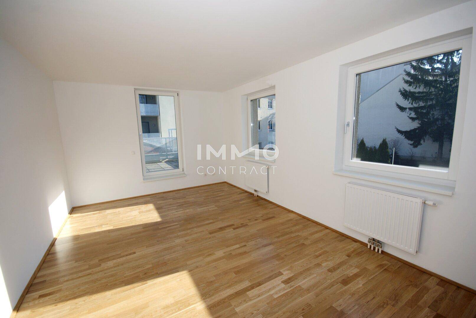 Heller Raum mit Holzboden, weißen Wänden und großen Fenstern, die viel Tageslicht hereinlassen.
