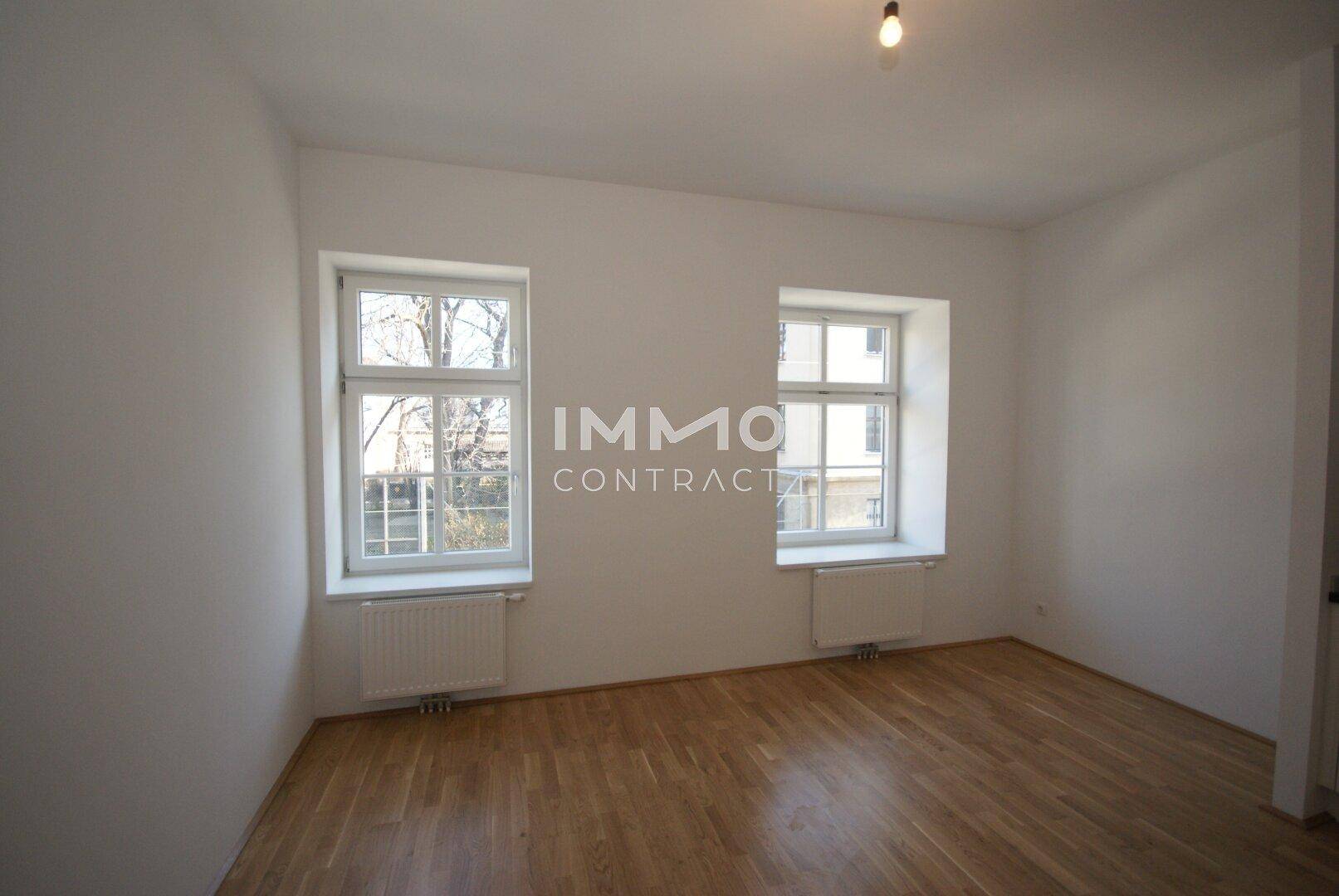 Geräumiger Raum mit Holzboden, weißen Wänden und zwei klassischen Fenstern, die Tageslicht spenden.
