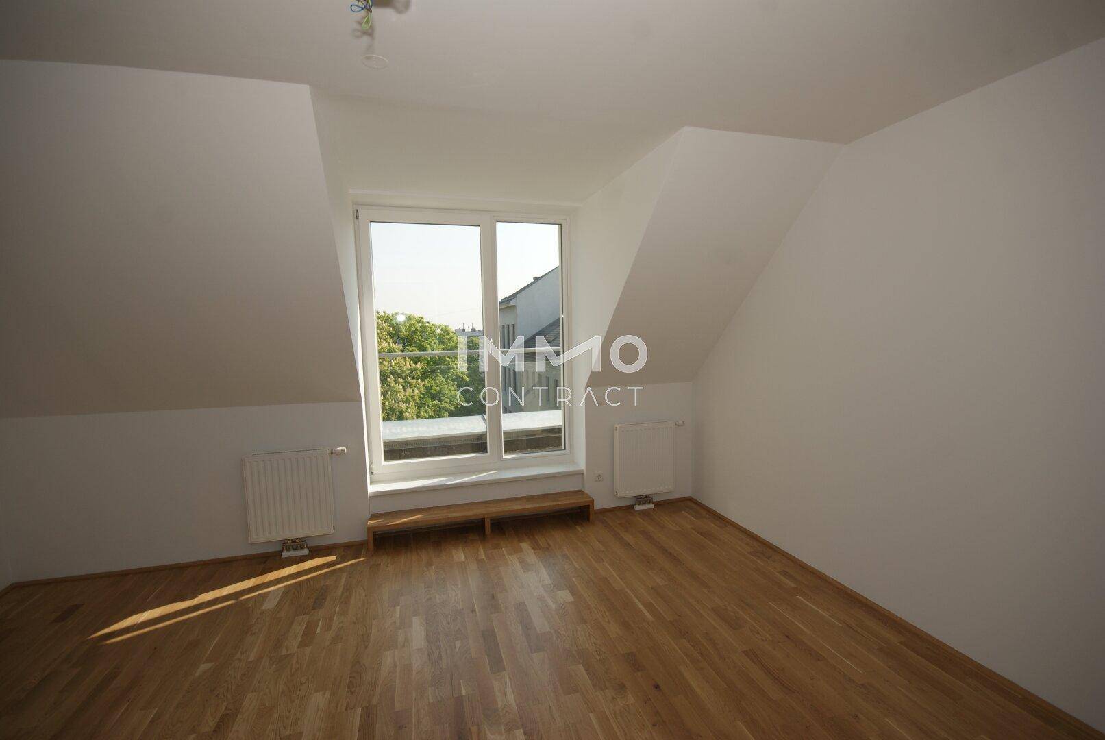 Heller Raum mit Holzboden, Dachschräge und einem großen Fenster, das viel Sonnenlicht einfängt.