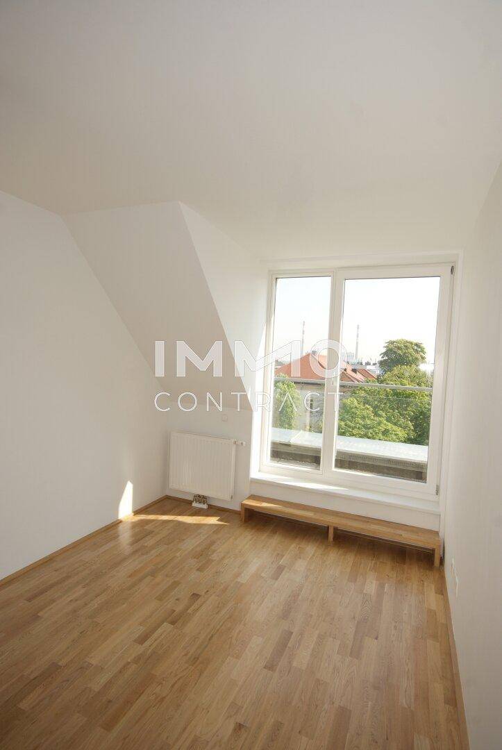 Lichtdurchfluteter Raum mit Holzboden, Dachschräge und großem Fenster mit Blick ins Grüne.