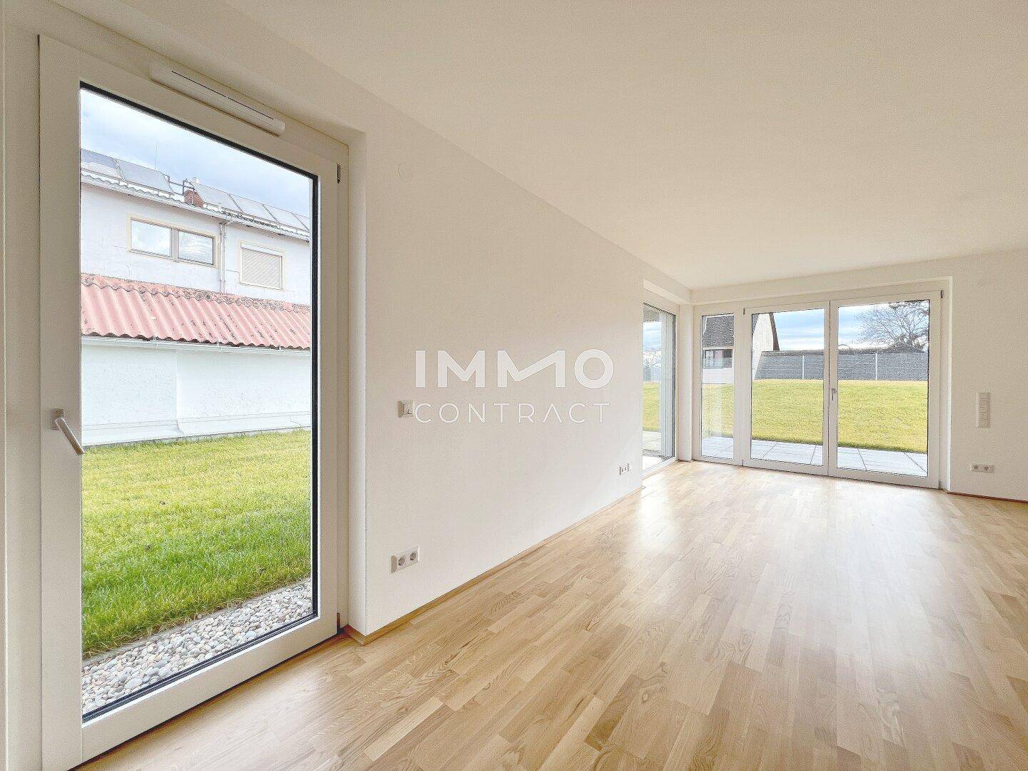 Heller, leerer Raum mit Holzparkettboden und großen Fenstern, die Zugang zum Garten bieten.