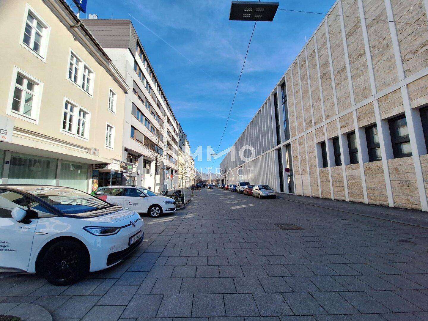 Breite Stadtstraße mit gepflastertem Gehweg, modernen Gebäuden und geparkten Autos unter blauem Himmel.