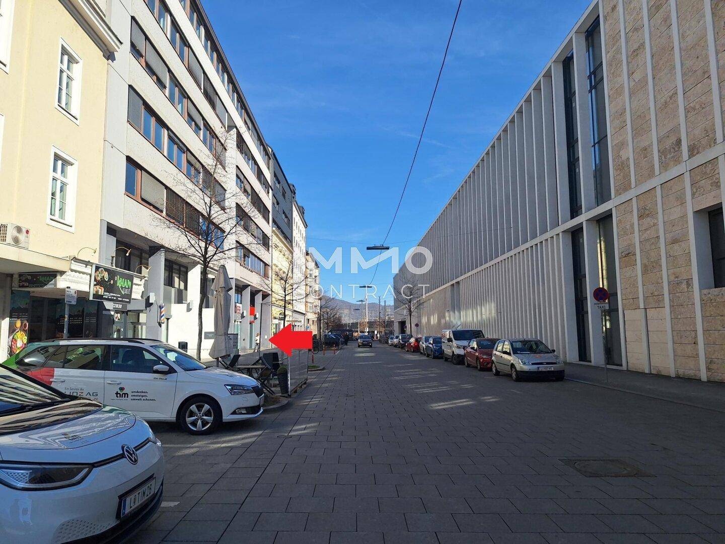 Stadtstraße mit geparkten Autos, modernen Gebäuden und einem belebten Gehweg unter klarem Himmel.