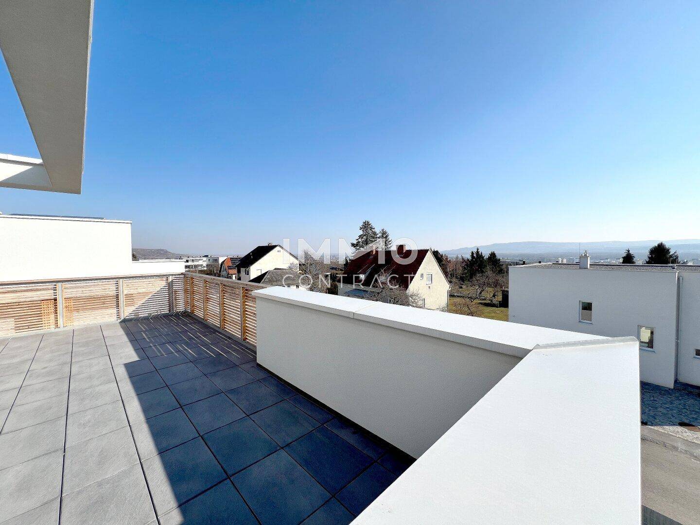 Sonniger Terrassenbereich mit moderner Architektur, Fliesenboden und Blick auf die umliegende Bebauung.