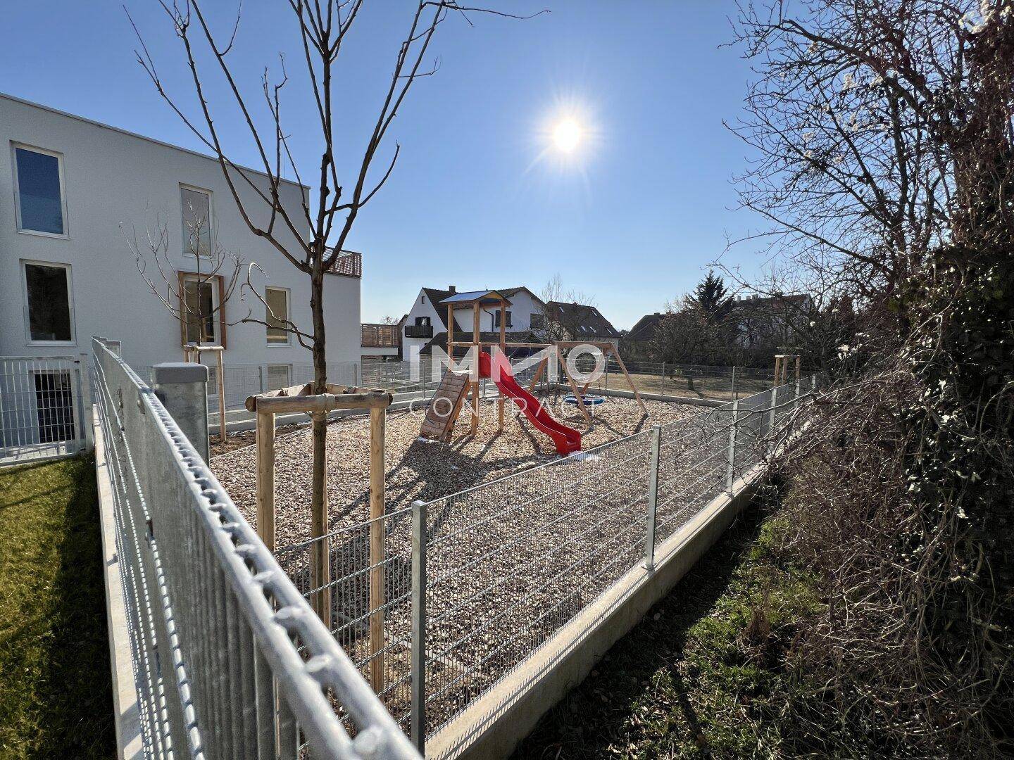 Außenbereich mit einem umzäunten Kinderspielplatz, ausgestattet mit Rutsche und Klettergerüst.