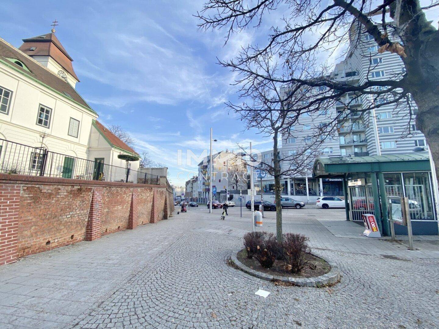 Stadtansicht mit Kopfsteinpflasterstraße, Gebäuden und Bäumen unter blauem Himmel.