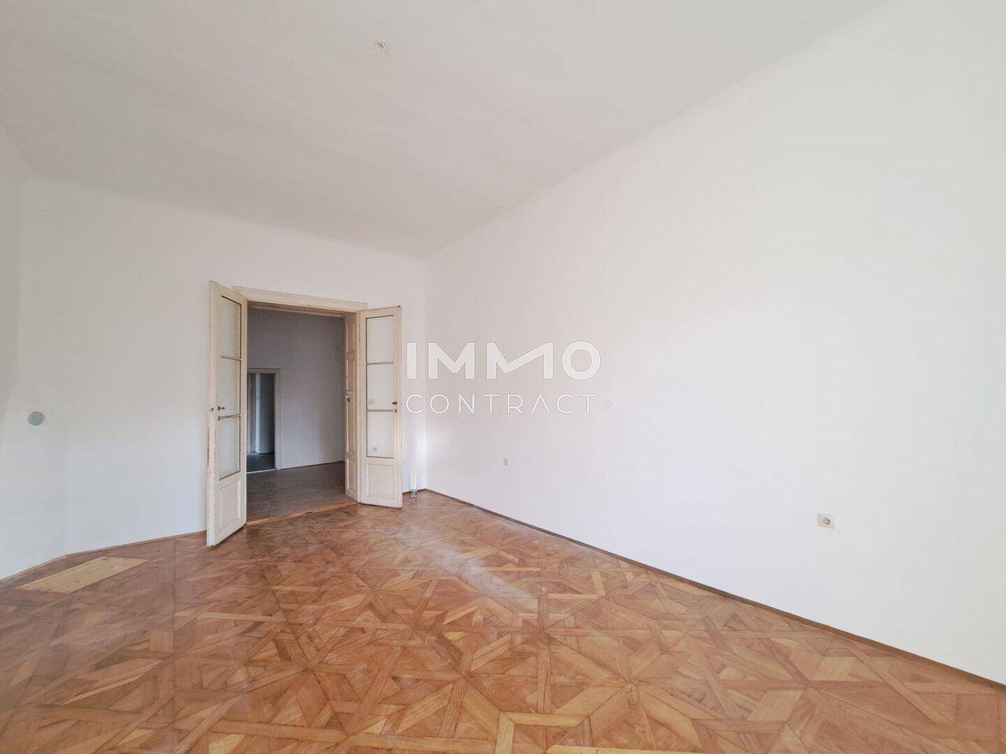 Geräumiger, leerer Innenraum mit weißen Wänden und einem klassischen Holzboden, der zu einem weiteren Zimmer führt.