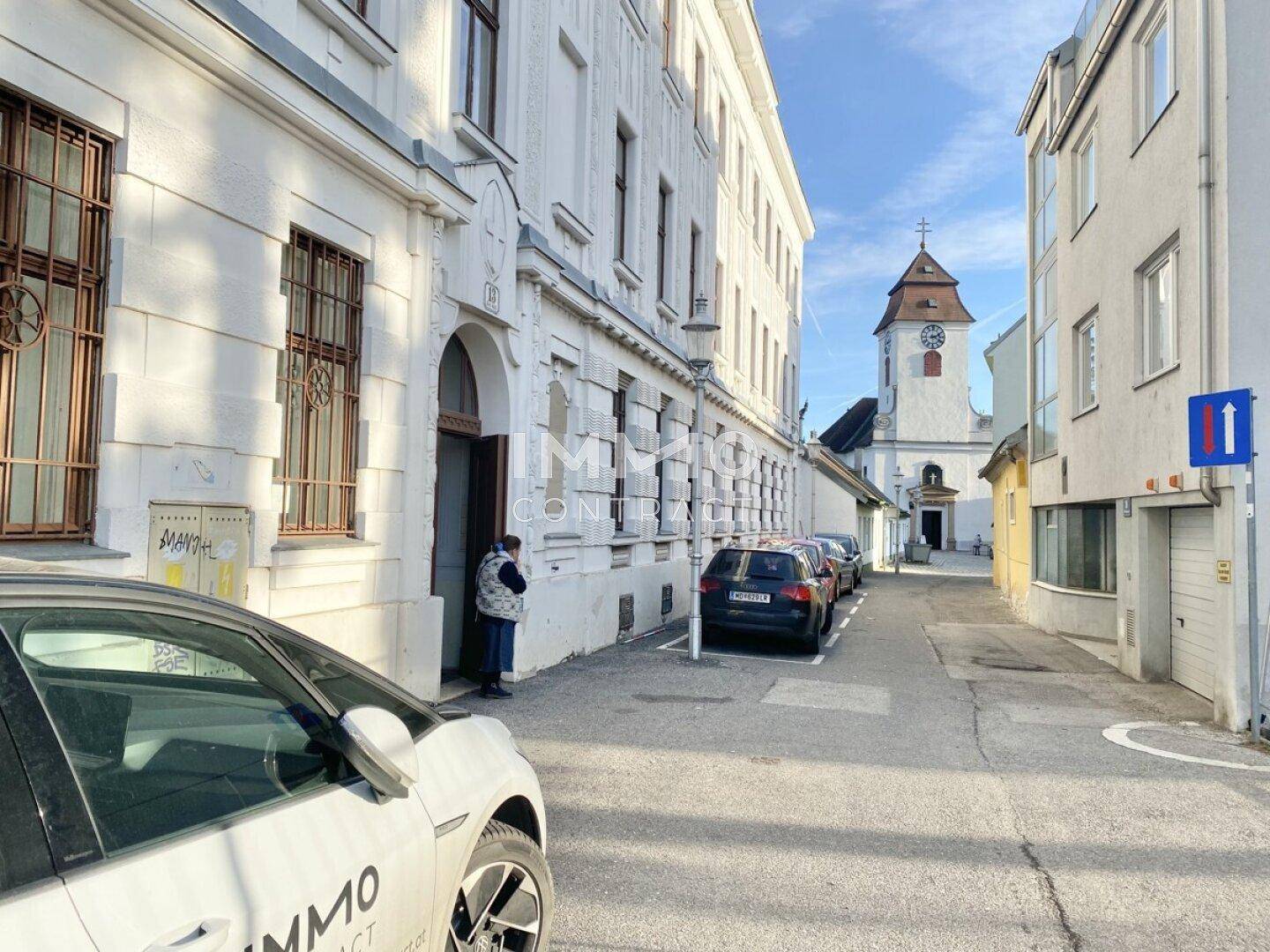 Historische Gebäude und eine Kirche in einer engen Gasse mit parkenden Autos.