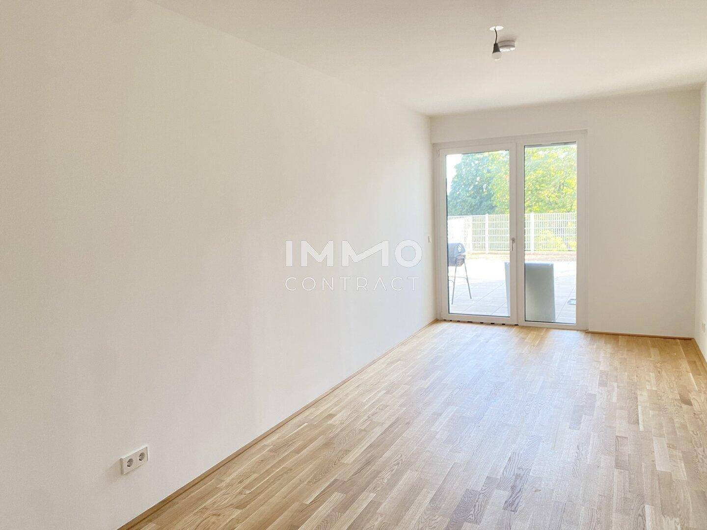 Heller, leerer Raum mit Holzboden und großen Fenstern, die auf eine Terrasse führen.