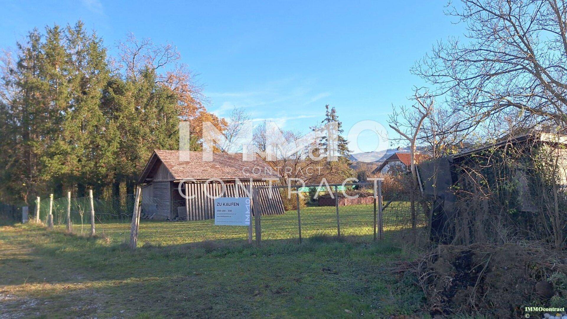 Gartenbereich mit Zaun und einem 'Zu Kaufen'-Schild vor einem Holzschuppen.