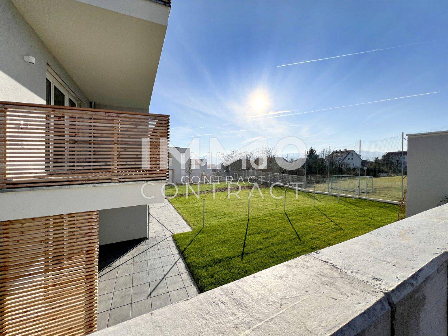 Sonniger Außenbereich mit Terrasse, Rasenfläche und Blick auf einen Sportplatz.