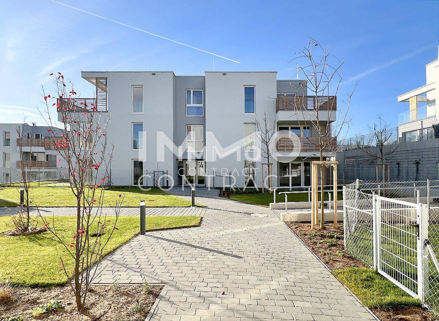 Moderne Wohnanlage mit gepflasterten Wegen, jungen Bäumen und einer hellen Fassade.